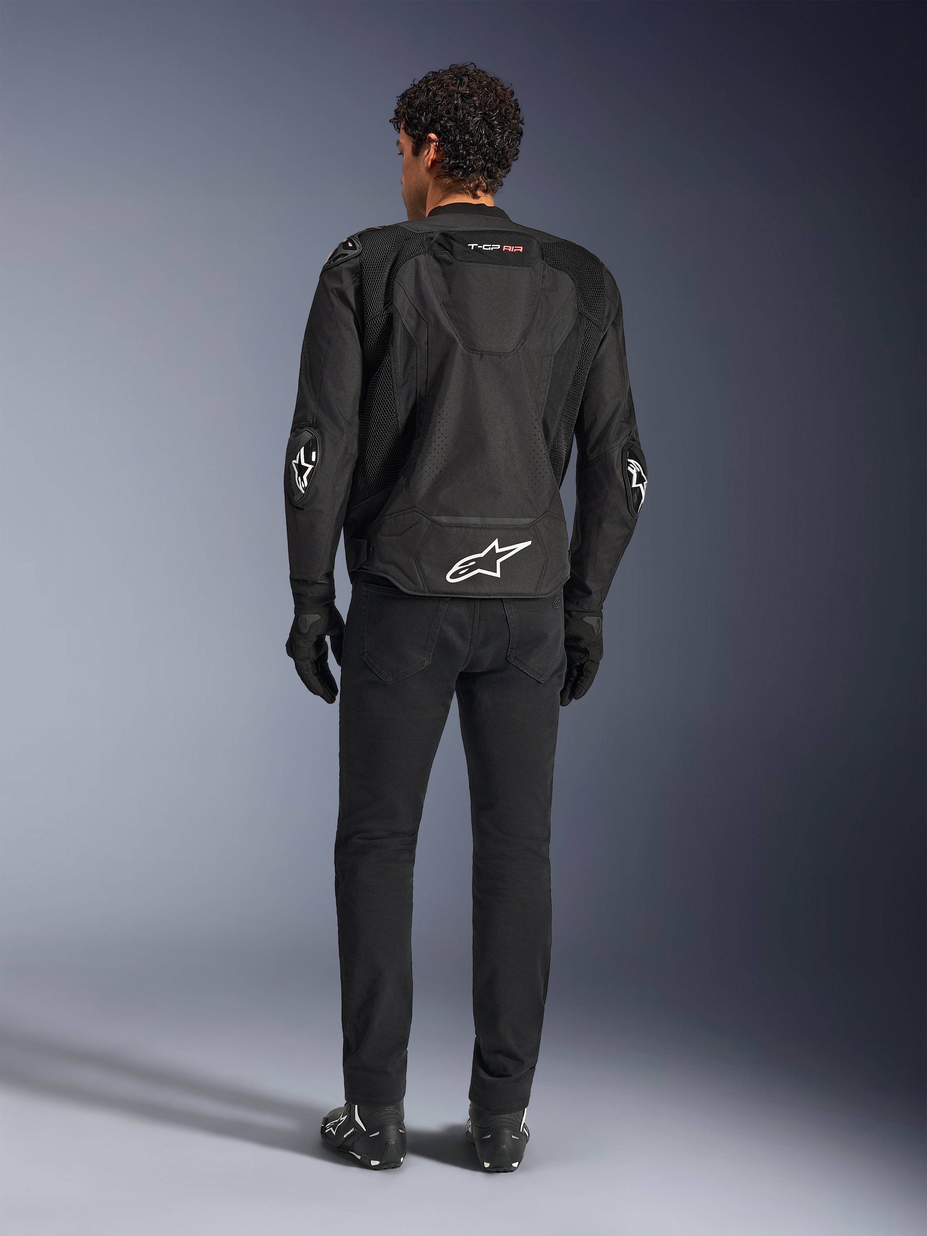 Casaco Alpinestars T-GP Air, preto, vista traseira no modelo, com painéis de ventilação em malha 3D e corcunda aerodinâmica, logótipo de estrela branco na zona lombar e cotovelos, usado com calças e luvas de motociclismo pretas, vestuário de condução desportiva.