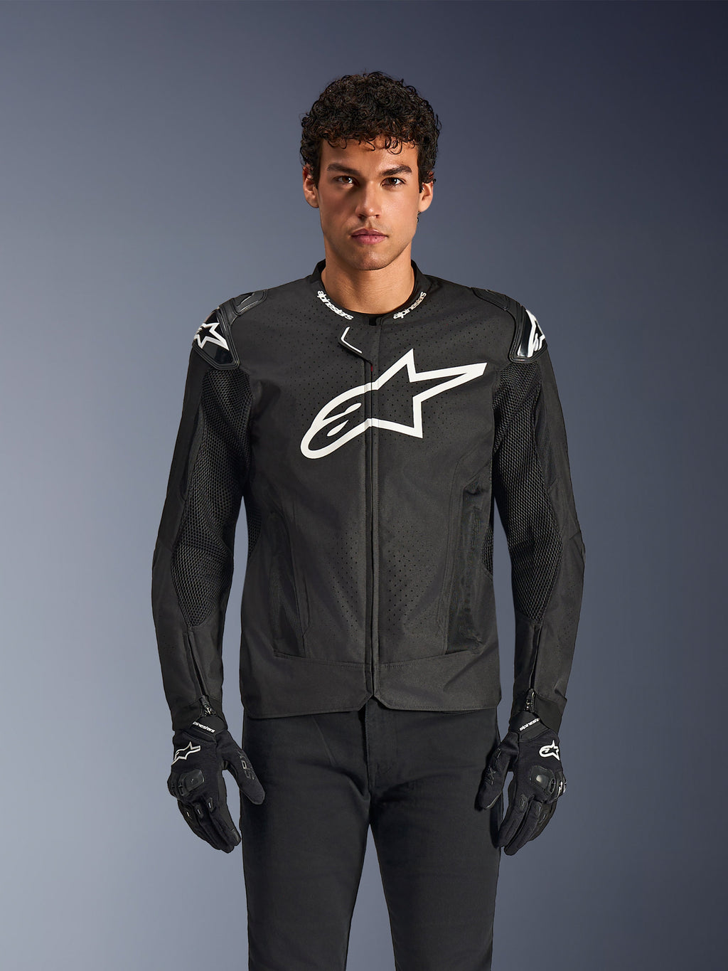 Alpinestars T-GP Air Jacket, casaco de motociclismo têxtil preto com um grande logótipo de estrela branco no peito, apresenta painéis de ventilação perfurados e malha 3D, deslizadores externos nos ombros, usado com luvas pretas e brancas, design desportivo elegante para condução em estrada com tempo quente