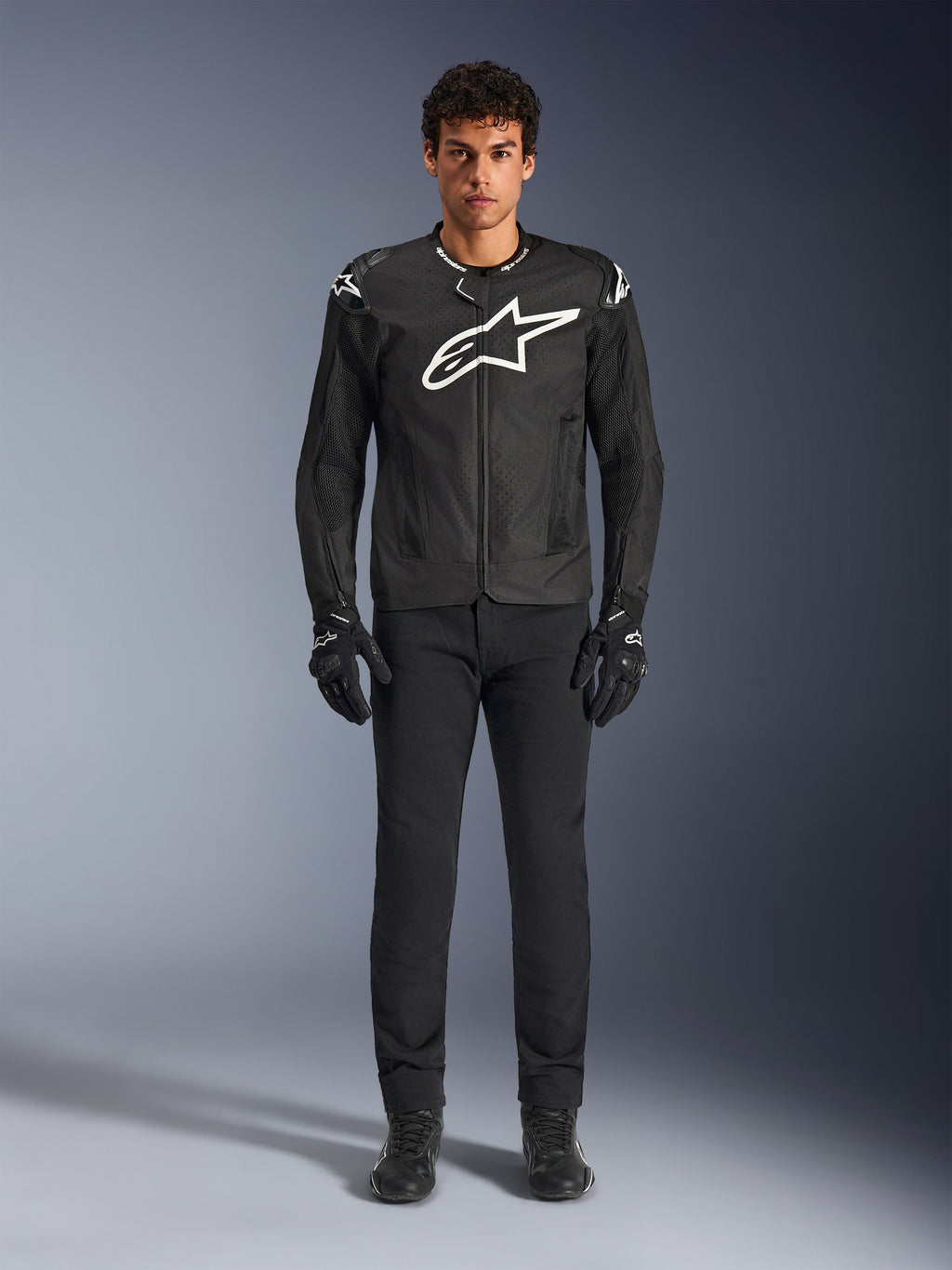 Casaco Alpinestars T-GP Air, casaco de motociclismo têxtil preto com um grande logótipo Astars branco no peito, usado por um modelo com calças de condução e luvas pretas, apresentando painéis perfurados e ventilação em malha para condução desportiva.