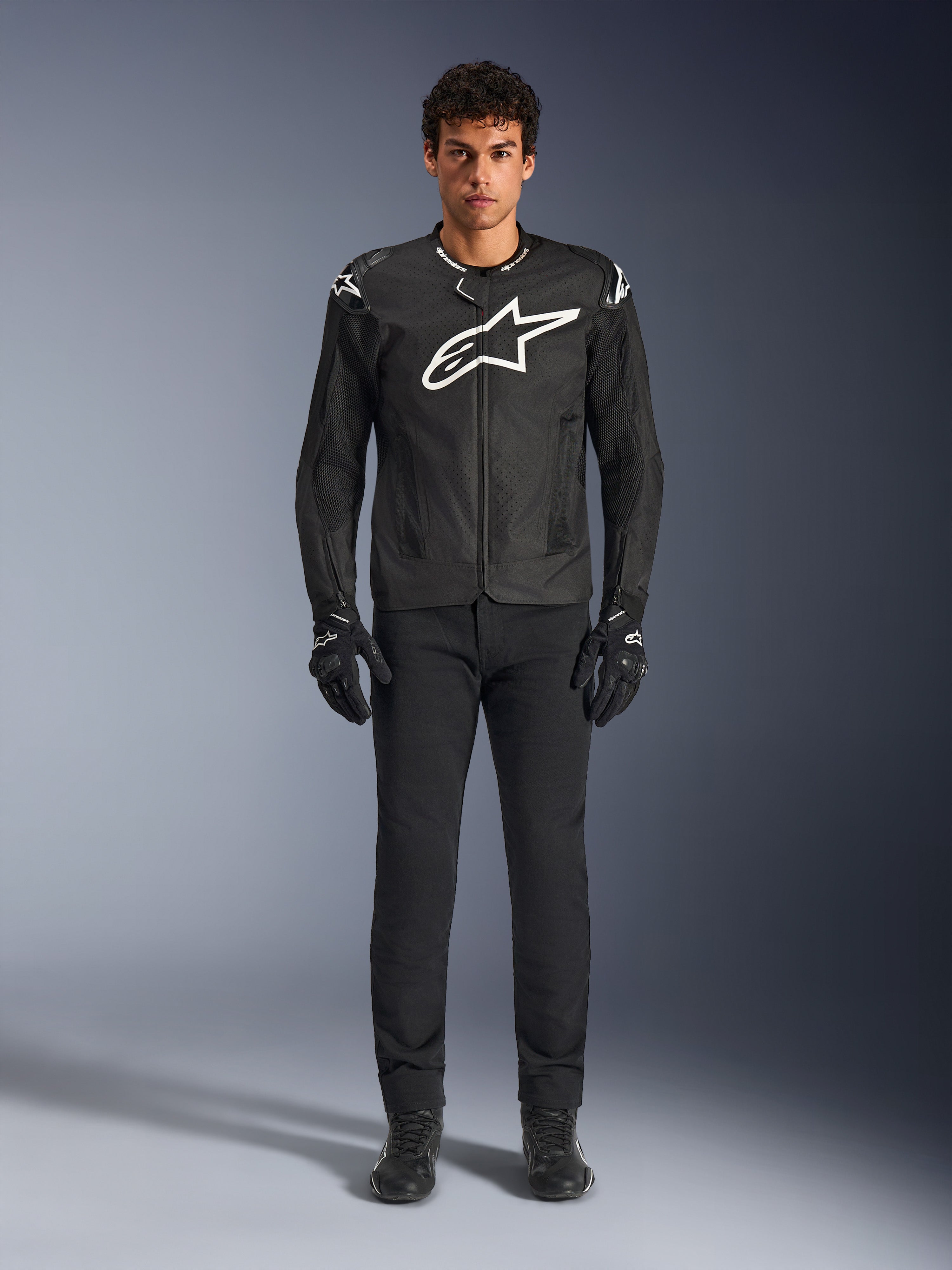 Casaco Alpinestars T-GP Air, casaco de motociclismo têxtil preto com um grande logótipo Astars branco no peito, usado por um modelo com calças de condução e luvas pretas, apresentando painéis perfurados e ventilação em malha para condução desportiva.