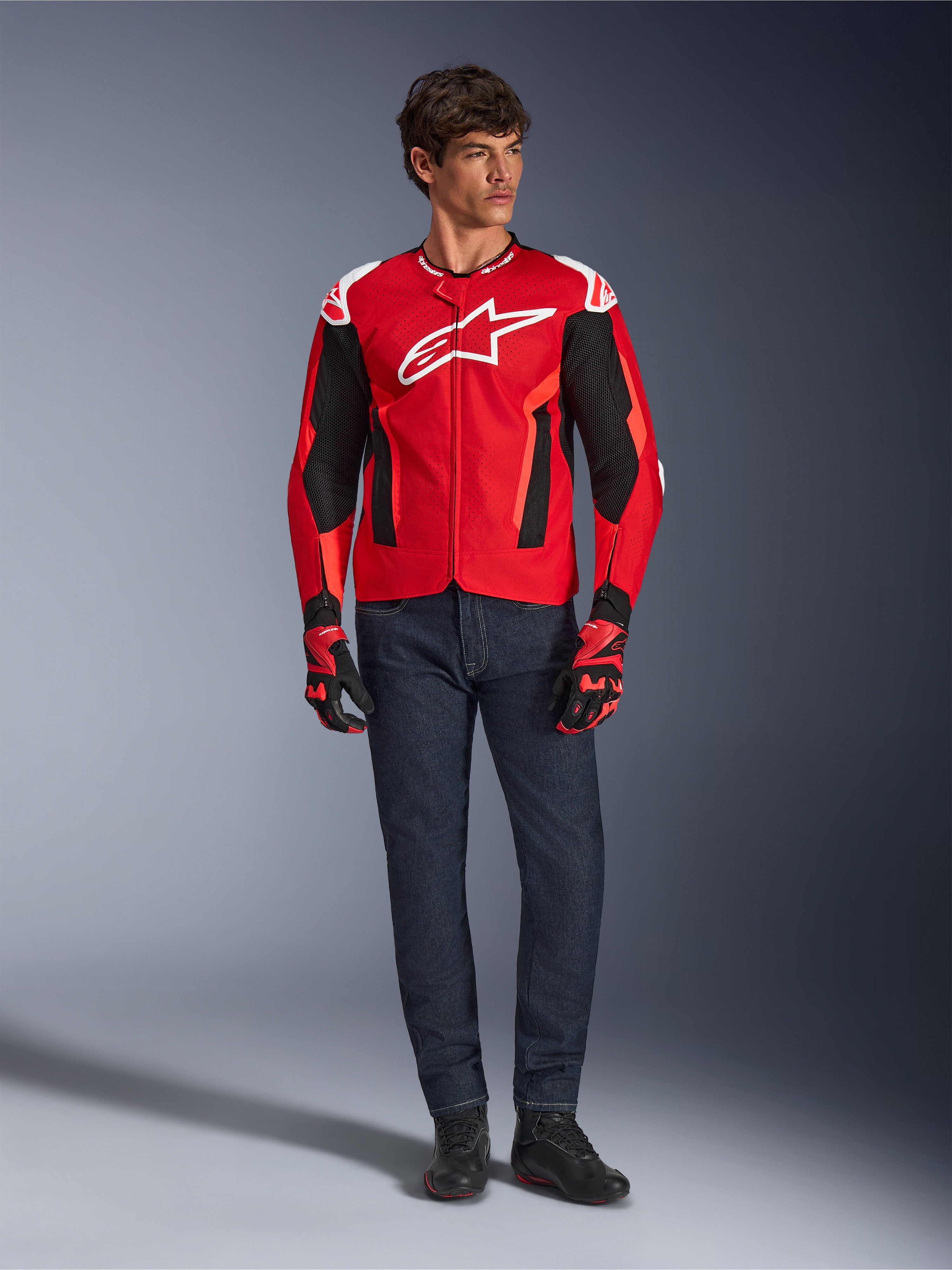 Alpinestars T-GP Air Jacket, Bright Red Red Fluo Black, male model wearing full motorcycle outfit with dark blue jeans and matching gloves, sport textile jacket featuring perforated mesh panels and white Astars logo, studio shot.