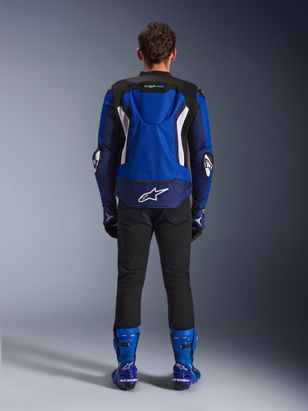 Casaco Alpinestars T-GP Air, Azul Brilhante, Azul Marinho e Branco, vista traseira com corcova aerodinâmica e painéis de ventilação em malha, logótipo Alpinestars branco na parte inferior das costas, usado com calças pretas e botas de corrida azuis