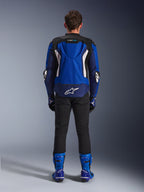 Casaco Alpinestars T-GP Air, Azul Brilhante, Azul Marinho e Branco, vista traseira com corcova aerodinâmica e painéis de ventilação em malha, logótipo Alpinestars branco na parte inferior das costas, usado com calças pretas e botas de corrida azuis