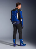 Casaco Alpinestars T-GP Air, casaco têxtil desportivo, azul brilhante, azul-marinho e branco, visto de trás num modelo a usar calças pretas e botas azuis e brancas a condizer, com logótipo Alpinestars branco na zona lombar, painéis de ventilação em malha e corcova aerodinâmica