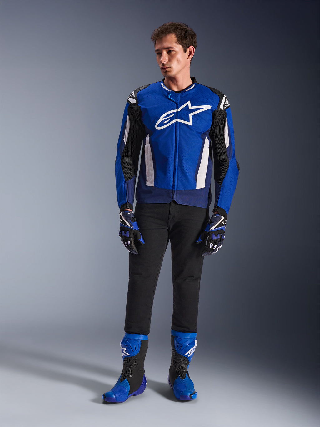 Modelo masculino a usar o Casaco Alpinestars T-GP Air em azul brilhante, azul marinho e branco, combinado com calças pretas, luvas de corrida em azul e azul marinho a condizer e botas de motociclismo em azul e preto, numa fotografia de estúdio que apresenta um equipamento completo de condução desportiva.