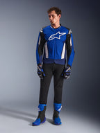 Modelo masculino a usar o Casaco Alpinestars T-GP Air em azul brilhante, azul marinho e branco, combinado com calças pretas, luvas de corrida em azul e azul marinho a condizer e botas de motociclismo em azul e preto, numa fotografia de estúdio que apresenta um equipamento completo de condução desportiva.