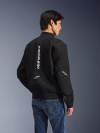 Casaco Alpinestars Flight Air, casaco de motociclismo preto, vista traseira com logótipo branco vertical grande e painéis de ventilação em malha, estilo bomber com bainha canelada, usado com calças de ganga azuis, equipamento de condução urbano contemporâneo