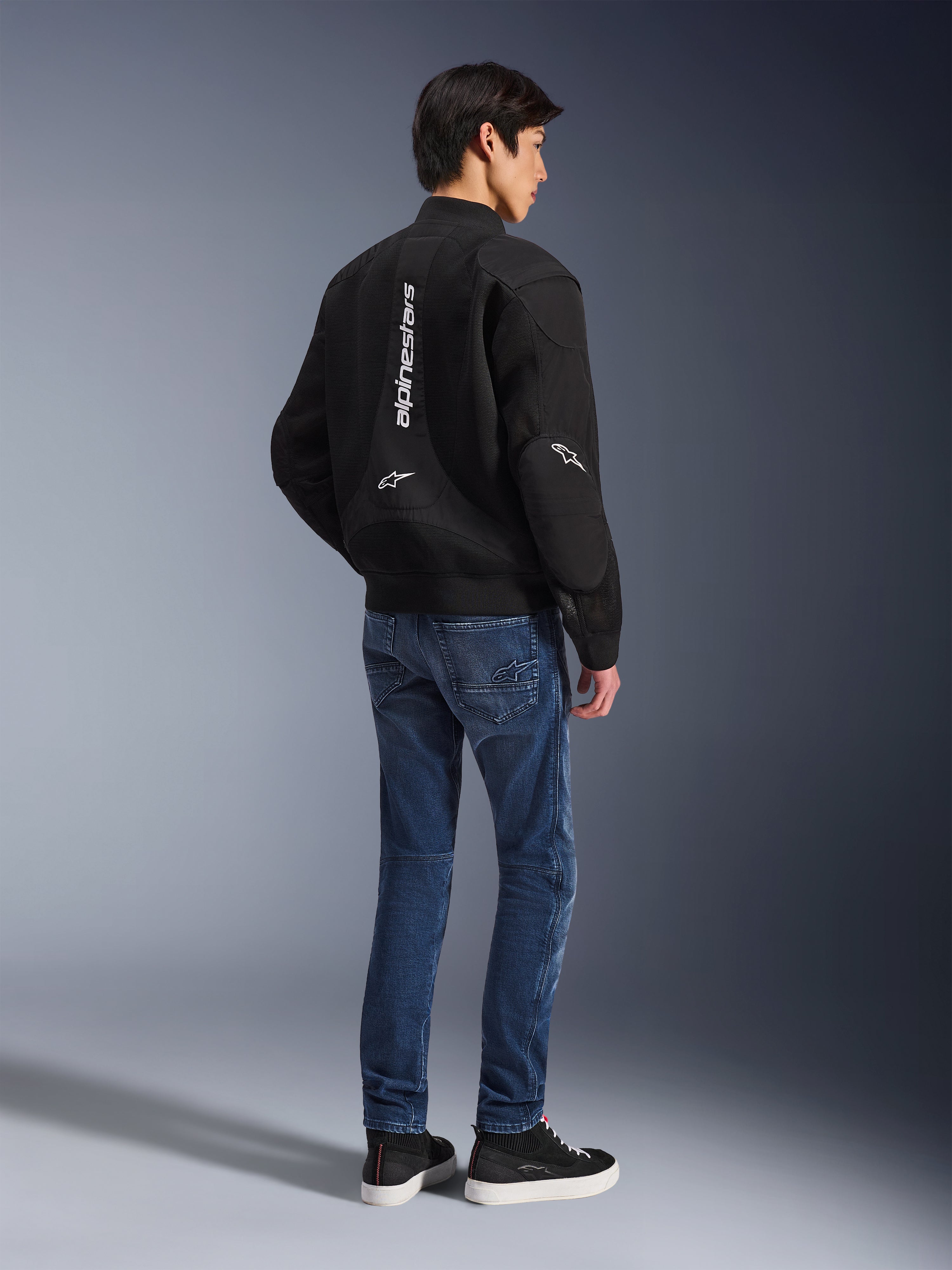 Modelo a usar o Casaco Alpinestars Flight Air em preto, visto de costas, combinado com calças de ganga de motociclismo azuis e sapatilhas pretas, com a marca Alpinestars vertical em branco e painéis de ventilação em malha 3D.