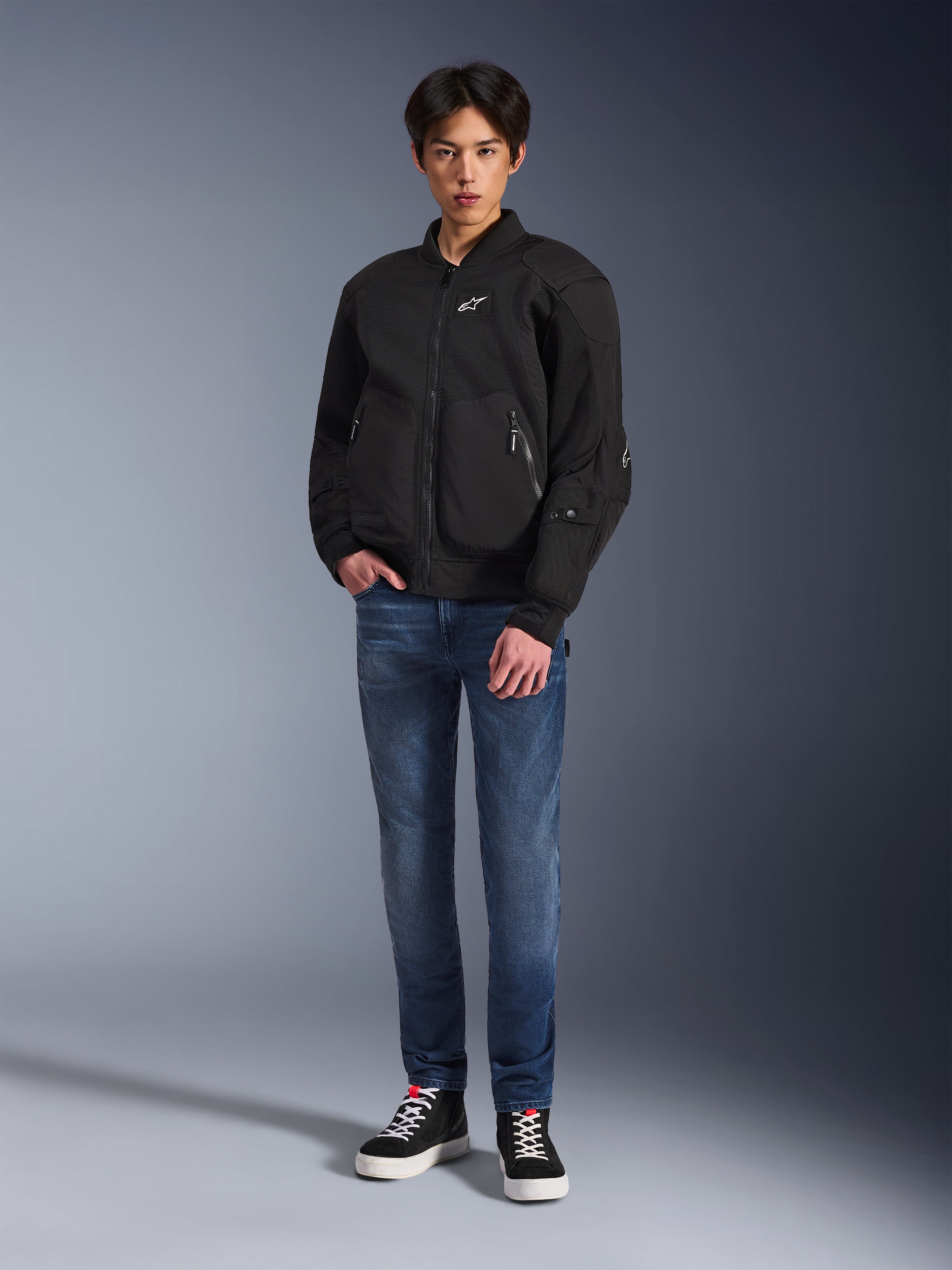 Modelo a usar o casaco Alpinestars Flight Air em preto, um casaco de motociclismo urbano de estilo bomber com painéis de malha 3D e fecho frontal, combinado com calças de ganga azuis e sapatilhas pretas, vestuário de condução contemporâneo de inspiração urbana.