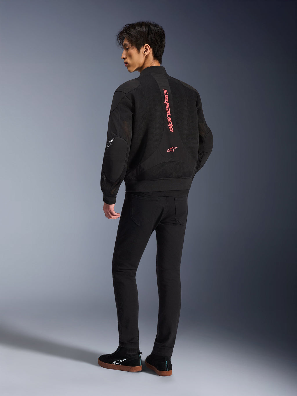 Male model wearing Alpinestars Flight Air Jacket in Black Bright Red, rear view showing vertical red branding on back and 3D mesh construction, paired with black pants and sneakers, urban motorcycle style