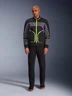 Modelo masculino a usar o Casaco Alpinestars Unite em Black Asphalt Violet, com base preta, painéis cinzentos e roxos, fechos e acabamentos em amarelo néon vibrante, combinado com calças e sapatos pretos, casaco de motociclismo urbano minimalista