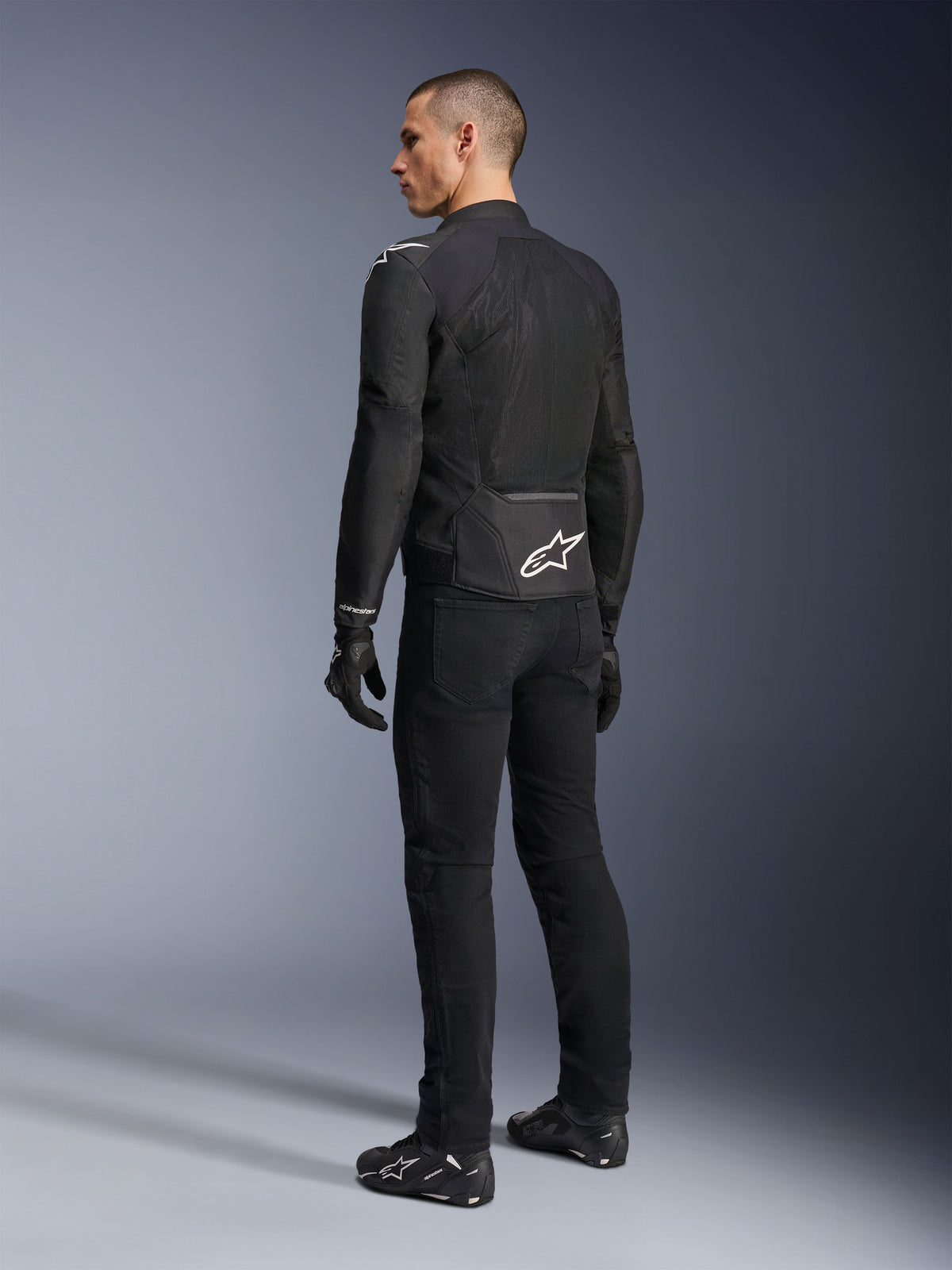 Casaco Alpinestars Viper Air V4, casaco de motociclismo em tecido preto, vista traseira num modelo masculino, com grandes painéis de malha tecnológica para fluxo de ar e um logótipo de estrela branco na parte inferior das costas, usado com calças e luvas de motociclismo pretas a condizer.