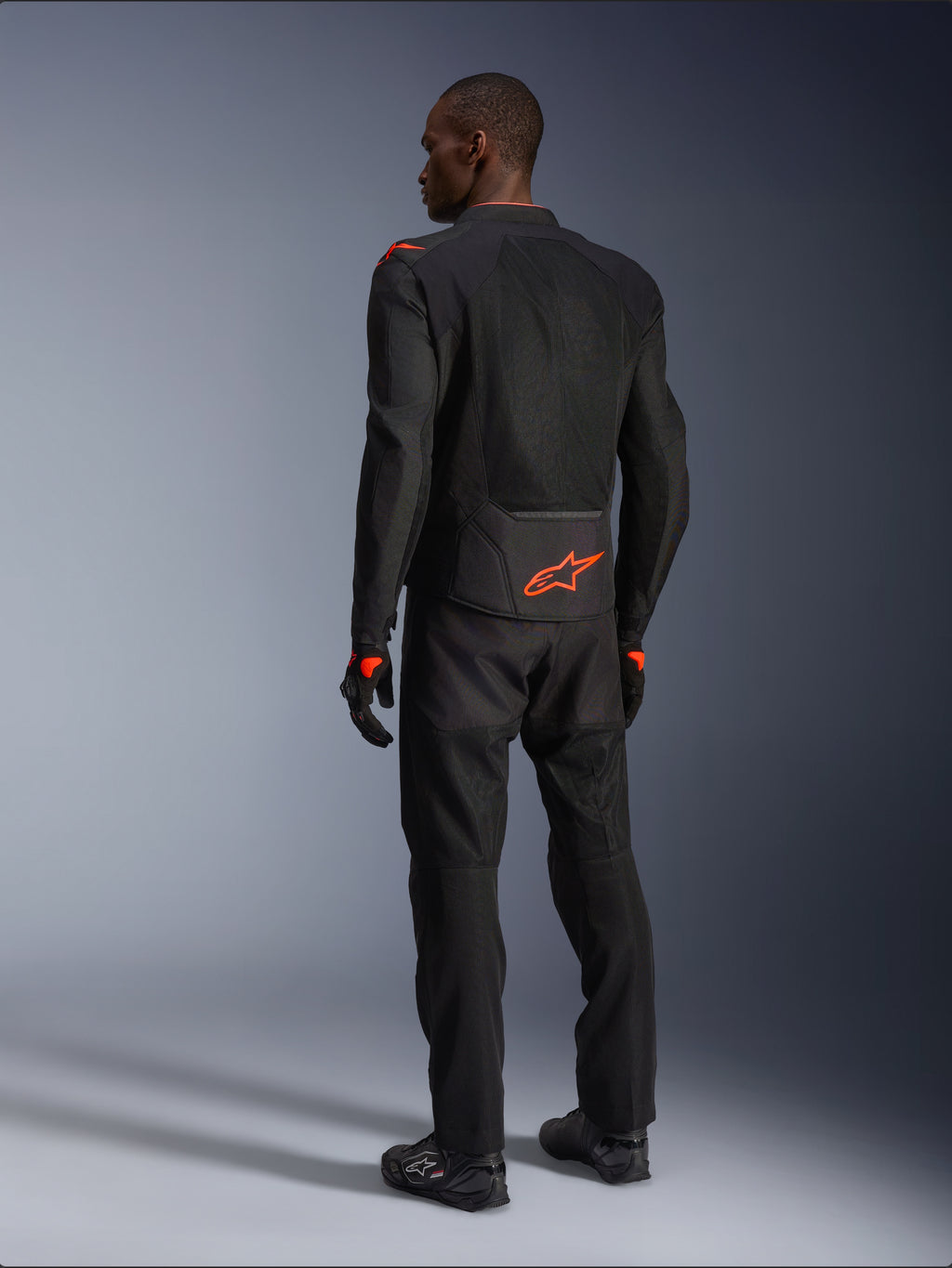 Male rider from the back wearing the Alpinestars Viper Air V4 Jacket, Black and Red Fluo, featuring an orange Astars logo on the lower back and shoulders, mesh textile construction, styled with matching black motorcycle pants and boots.