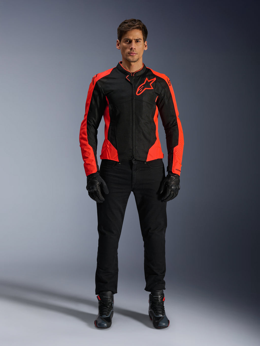 Modelo masculino a usar o Casaco Alpinestars Viper Air V4 em Vermelho Fluo e Preto, combinado com calças de motociclismo, luvas e botas pretas, apresentando painéis de malha respirável para ventilação e um logótipo de estrela proeminente no peito, vestuário desportivo para motociclismo de estrada