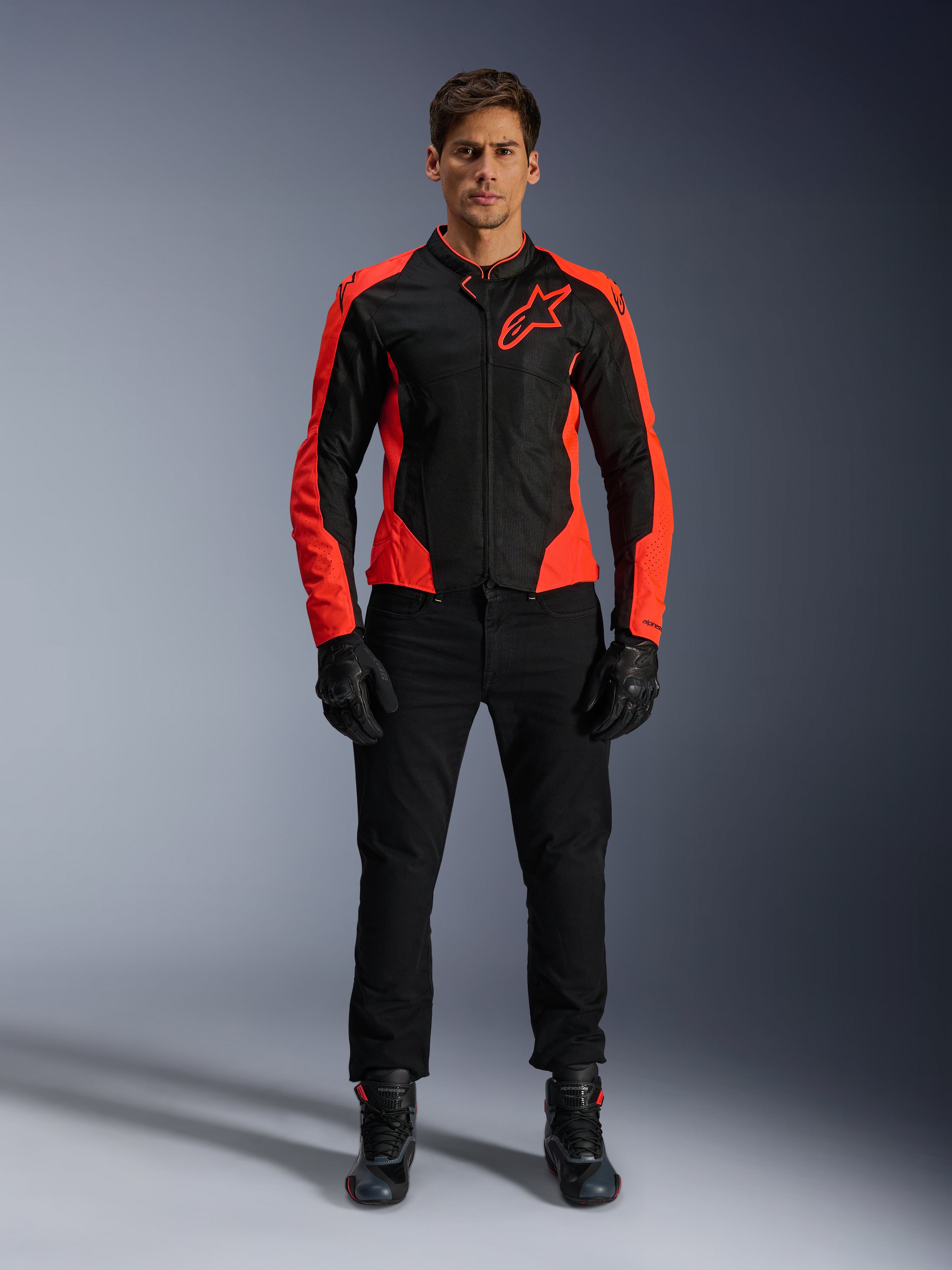Modelo masculino a usar o Casaco Alpinestars Viper Air V4 em Vermelho Fluo e Preto, combinado com calças de motociclismo, luvas e botas pretas, apresentando painéis de malha respirável para ventilação e um logótipo de estrela proeminente no peito, vestuário desportivo para motociclismo de estrada