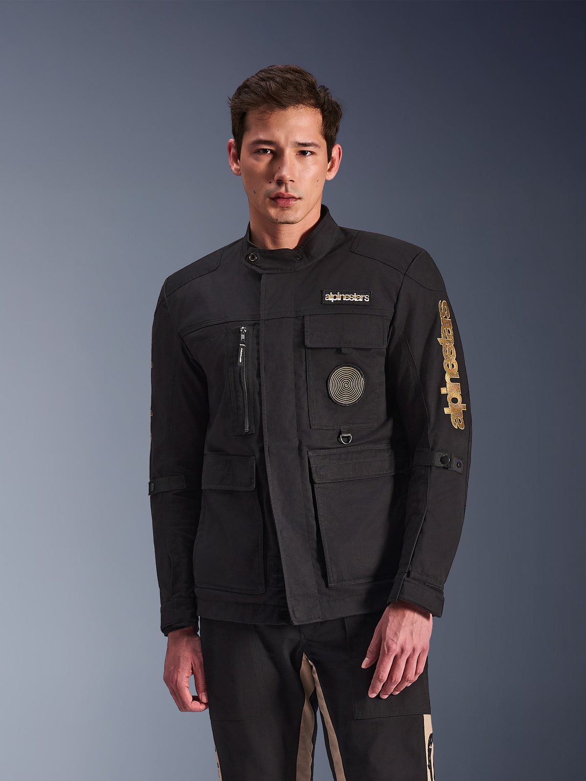 Alpinestars Dice Jacket, casaco de motociclista preto, usado por um modelo, com logótipos Alpinestars dourados, vários bolsos funcionais, emblema circular em espiral e estilo field jacket urbano