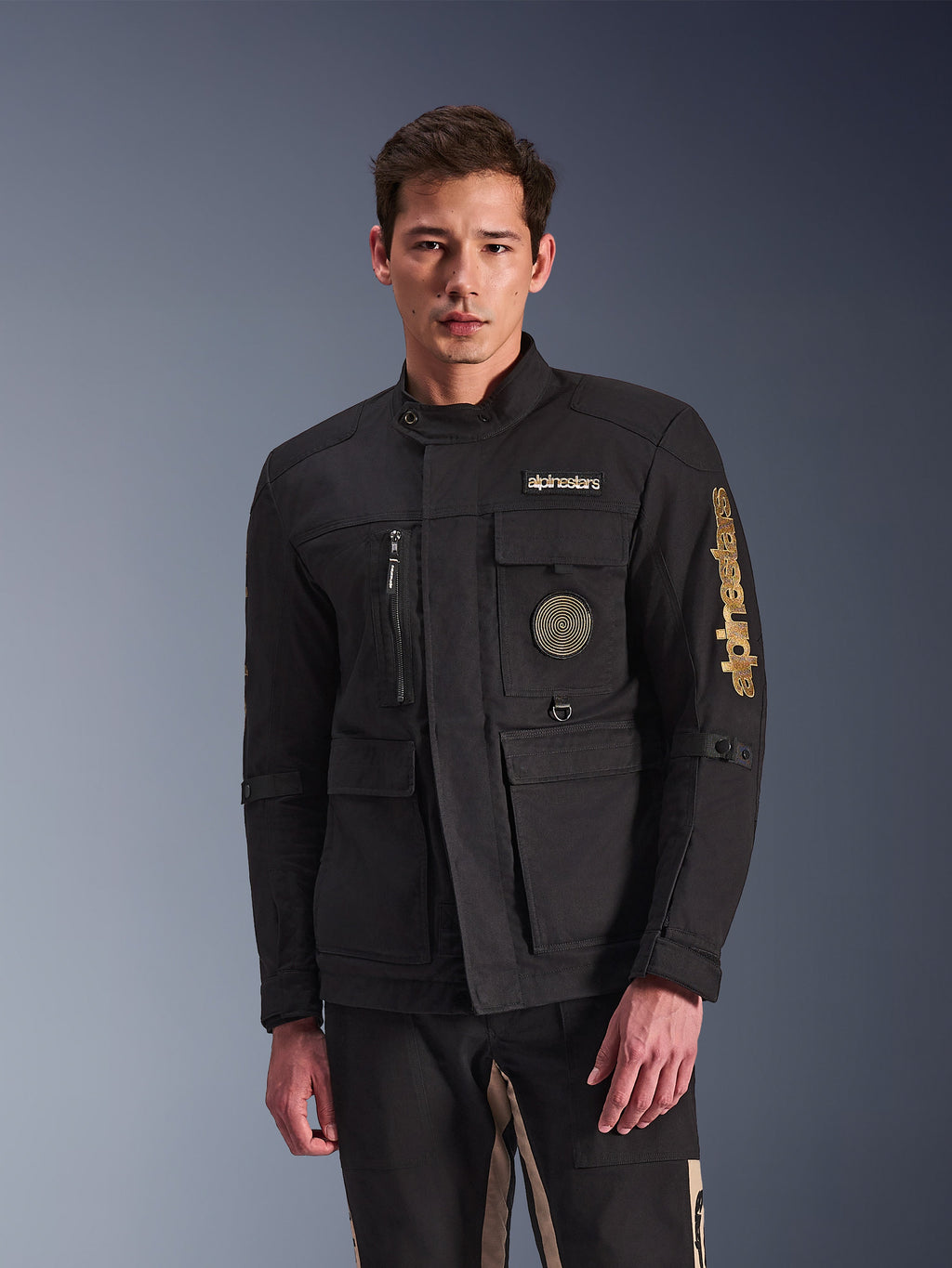 Alpinestars Dice Jacket, casaco de motociclista preto, usado por um modelo, com logótipos Alpinestars dourados, vários bolsos funcionais, emblema circular em espiral e estilo field jacket urbano