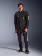 Modelo masculino a usar o Casaco Dice da Alpinestars, casaco de motociclista urbano preto com estilo vintage de casaco de campo, quatro bolsos frontais funcionais, marca Alpinestars na manga e no peito, combinado com calças pretas e beges, equipamento de condução urbana moderno