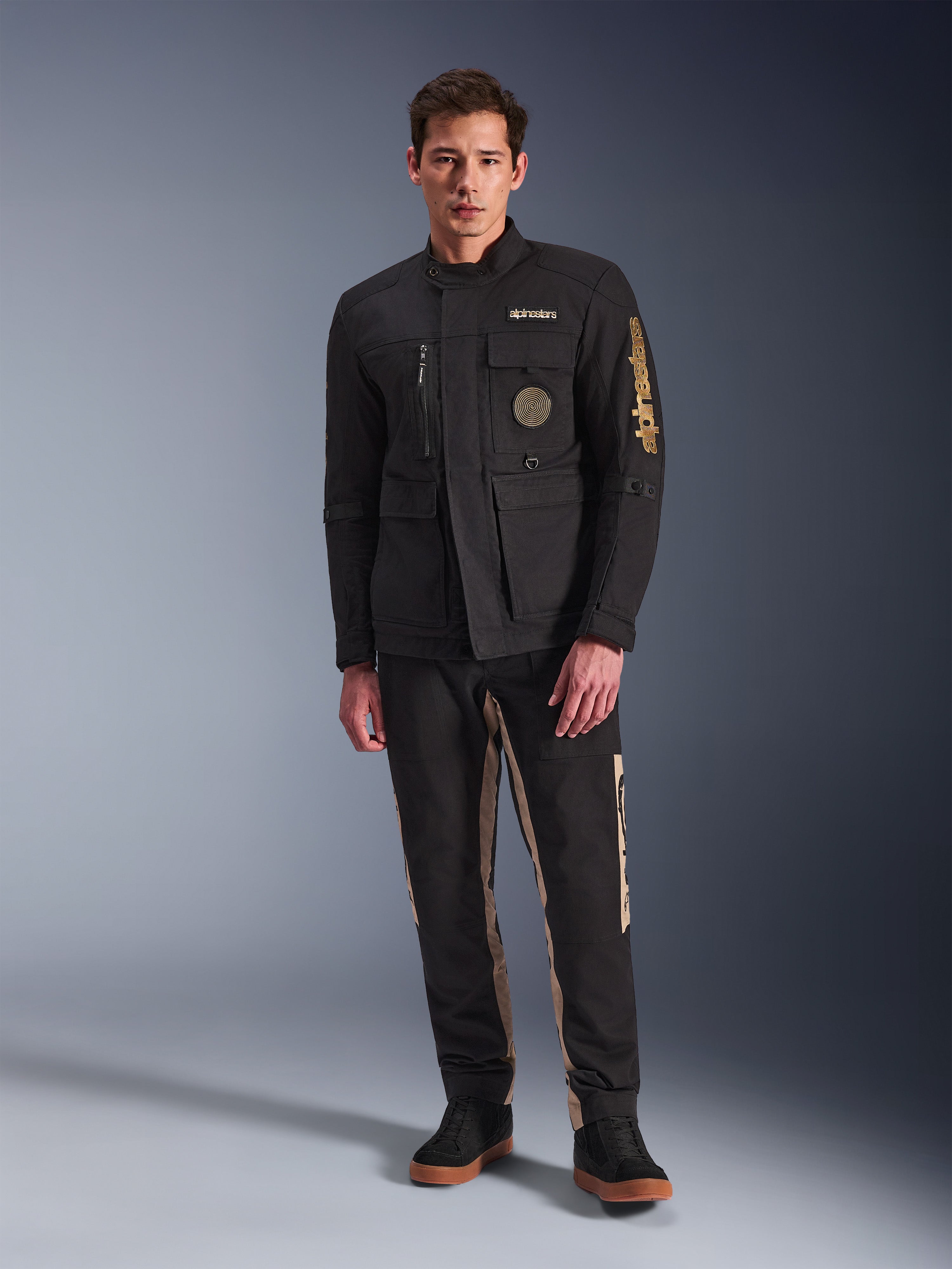 Modelo masculino a usar o Casaco Dice da Alpinestars, casaco de motociclista urbano preto com estilo vintage de casaco de campo, quatro bolsos frontais funcionais, marca Alpinestars na manga e no peito, combinado com calças pretas e beges, equipamento de condução urbana moderno