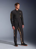 Modelo masculino a usar o Casaco Alpinestars Dice em preto, casaco de motociclista urbano com estética vintage de casaco de campo, com múltiplos bolsos utilitários e logótipo dourado na manga, combinado com calças pretas e beges a condizer e sapatilhas pretas