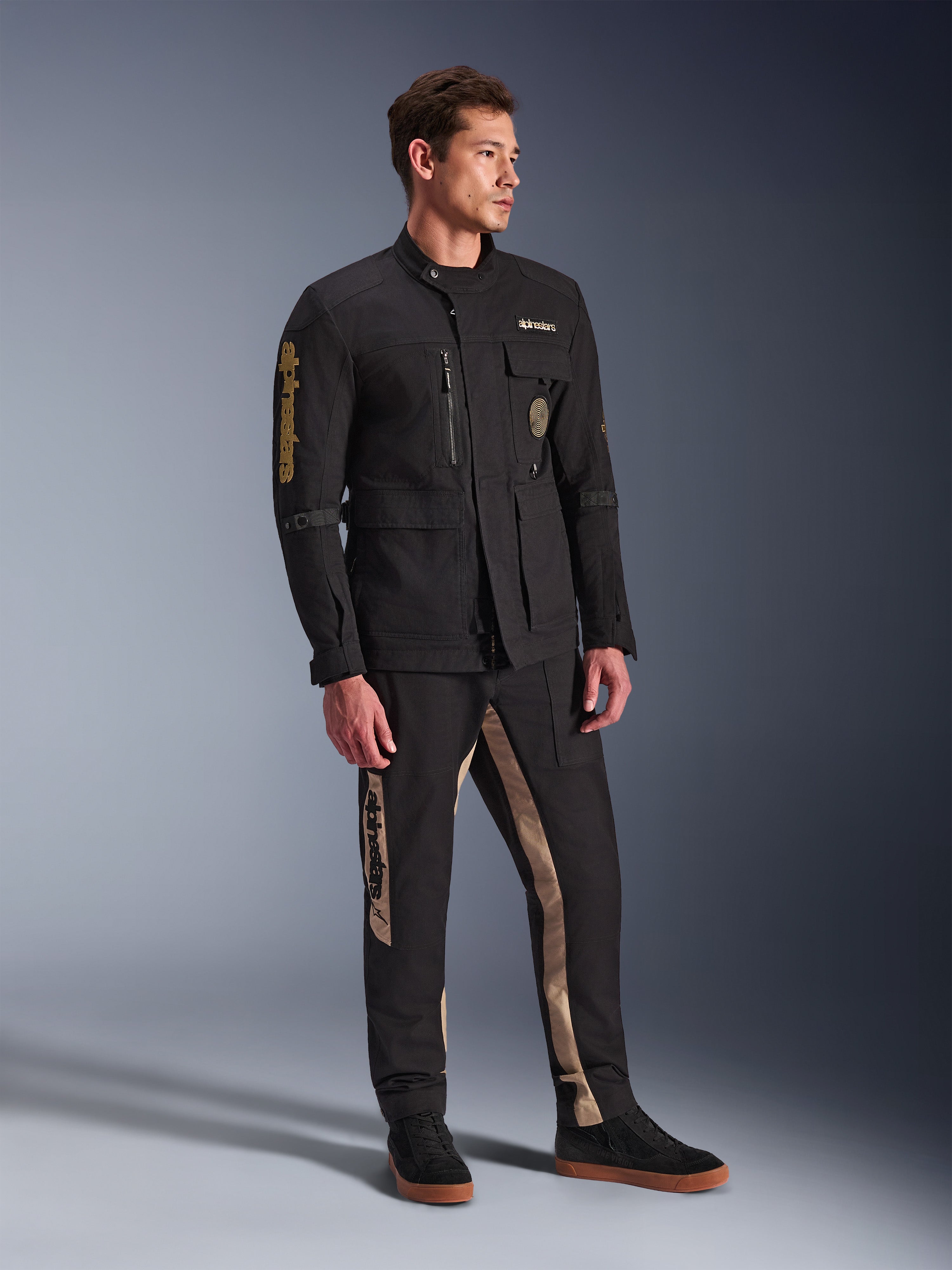 Modelo masculino a usar o Casaco Alpinestars Dice em preto, casaco de motociclista urbano com estética vintage de casaco de campo, com múltiplos bolsos utilitários e logótipo dourado na manga, combinado com calças pretas e beges a condizer e sapatilhas pretas