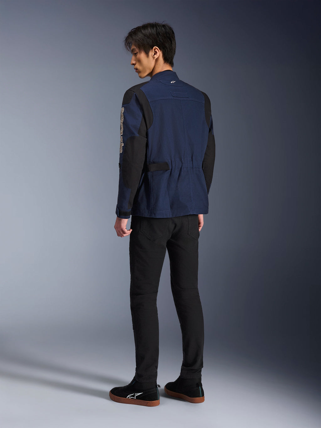 Male model viewed from the back wearing the Alpinestars Dice Jacket in Dark Blue and Black, paired with black pants and black sneakers, urban motorcycle field jacket featuring box-pleated shoulders and adjustable waist tabs.