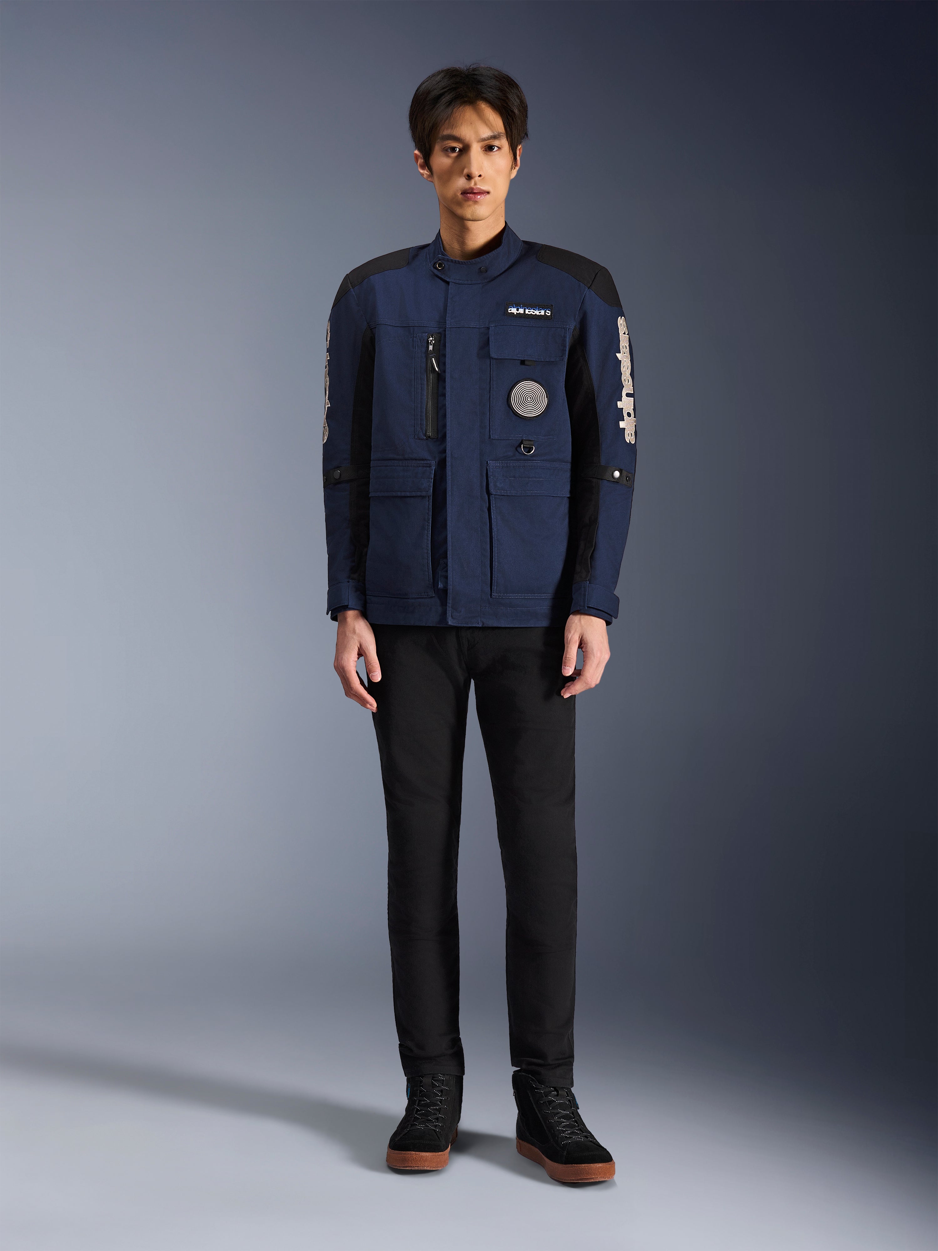Alpinestars Dice Jacket, Urban Motorcycle Jacket, Dark Blue and Black, worn by a model with black pants and black shoes, modern field jacket design with multiple pockets, vertical chest zipper, and white Alpinestars branding on the sleeves.