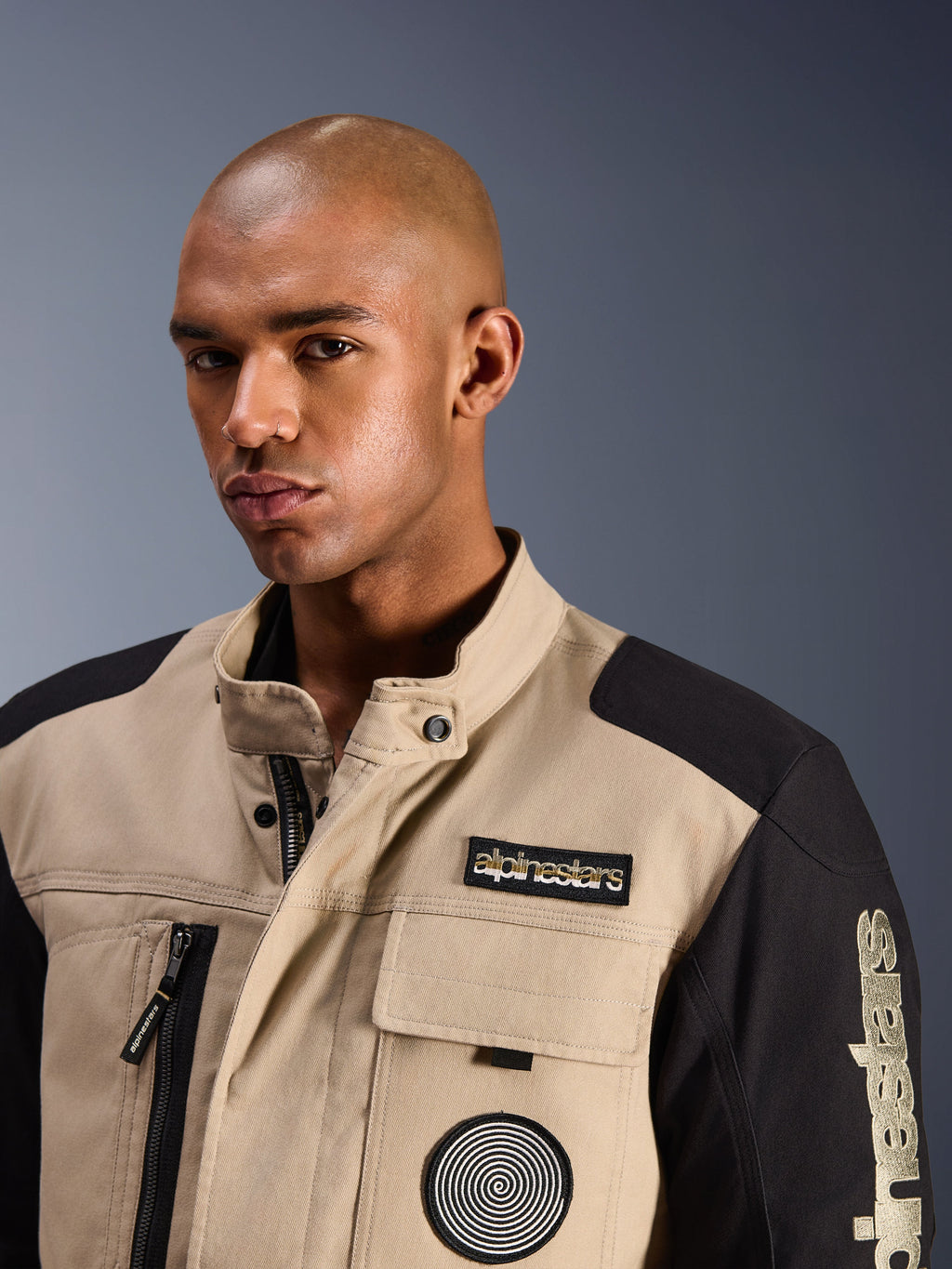 Modelo masculino a usar o Casaco Alpinestars Dice em Aluminum Black, casaco de motociclista urbano com painéis em blocos de cores bege e preto, com um patch bordado da Alpinestars no peito e logótipo em espiral, design robusto inspirado em vestuário de trabalho com gola alta