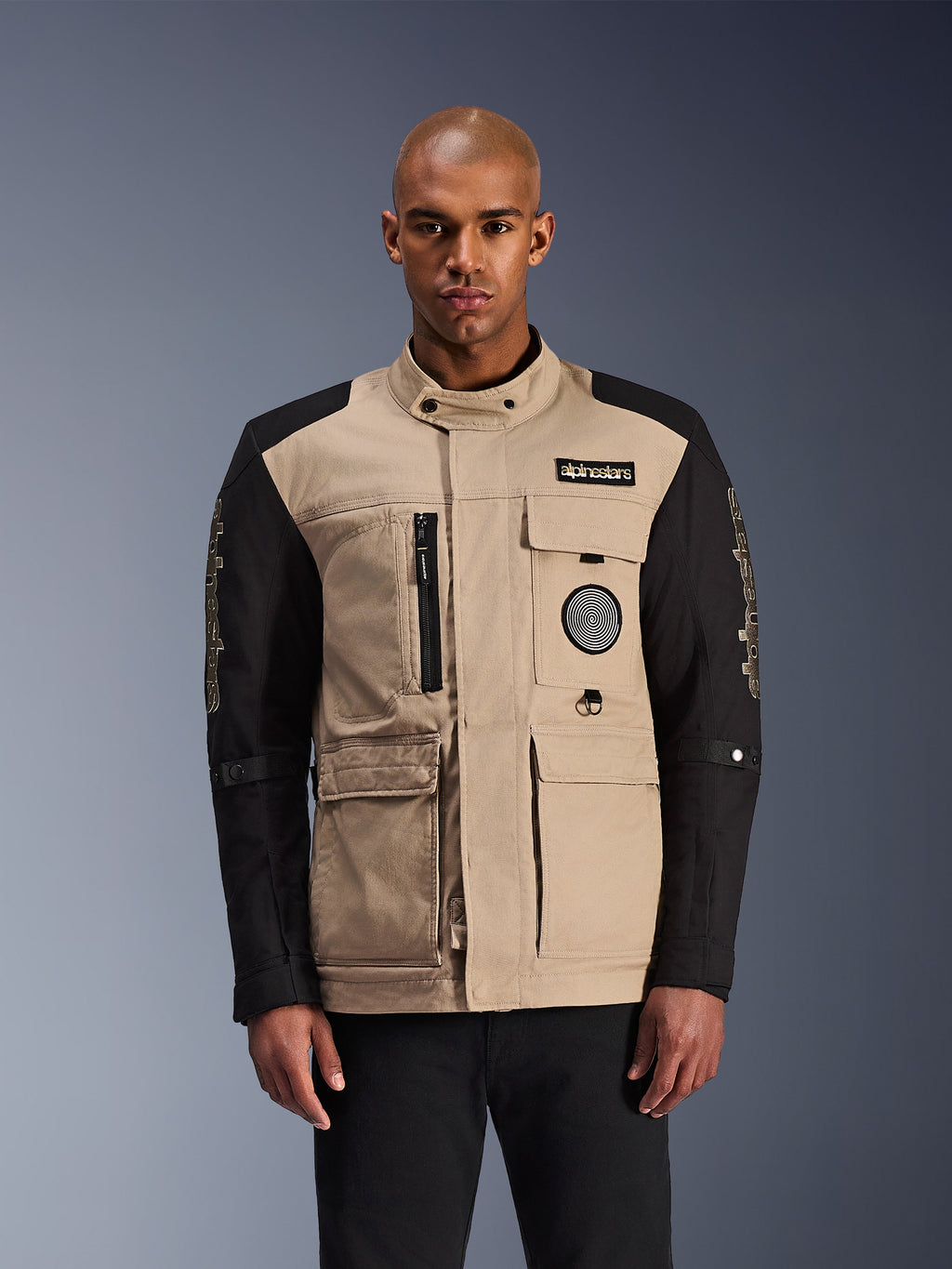 Alpinestars Dice Jacket, casaco de moto urbano, bege e preto, estética robusta de vestuário de trabalho com múltiplos bolsos utilitários e patches no peito, colarinho alto com fecho de botão, logótipos marcantes nas mangas, vestido por um modelo masculino