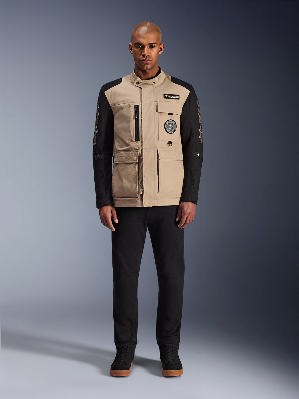 Casaco Alpinestars Dice, casaco de motociclismo urbano, corpo bege alumínio com mangas pretas, silhueta inspirada em vestuário de trabalho com quatro bolsos utilitários frontais, usado com calças pretas e sapatilhas de cano alto pretas, equipamento de condução robusto e urbano