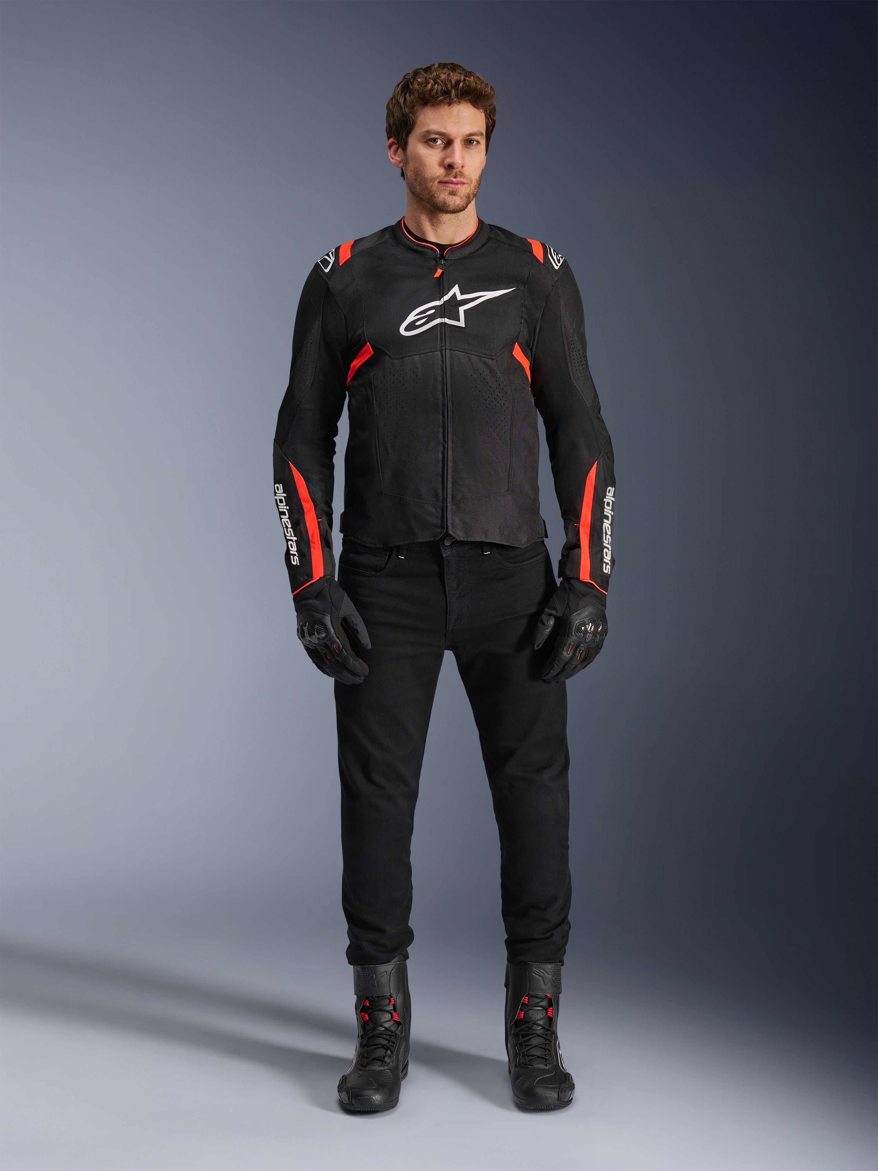 Modelo masculino a usar o casaco Alpinestars T-SPS Air v2, casaco de motociclismo em tecido preto com detalhes em vermelho fluo, logótipo branco no peito, calças, luvas e botas pretas, estilo de condução desportiva urbana.