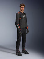 Modelo masculino a usar o Casaco Alpinestars T-Sps Air V2, preto com detalhes em vermelho fluorescente, combinado com calças de motociclismo pretas, luvas pretas e botas pretas, destacando o design desportivo com painéis de malha e logótipos Alpinestars brancos.