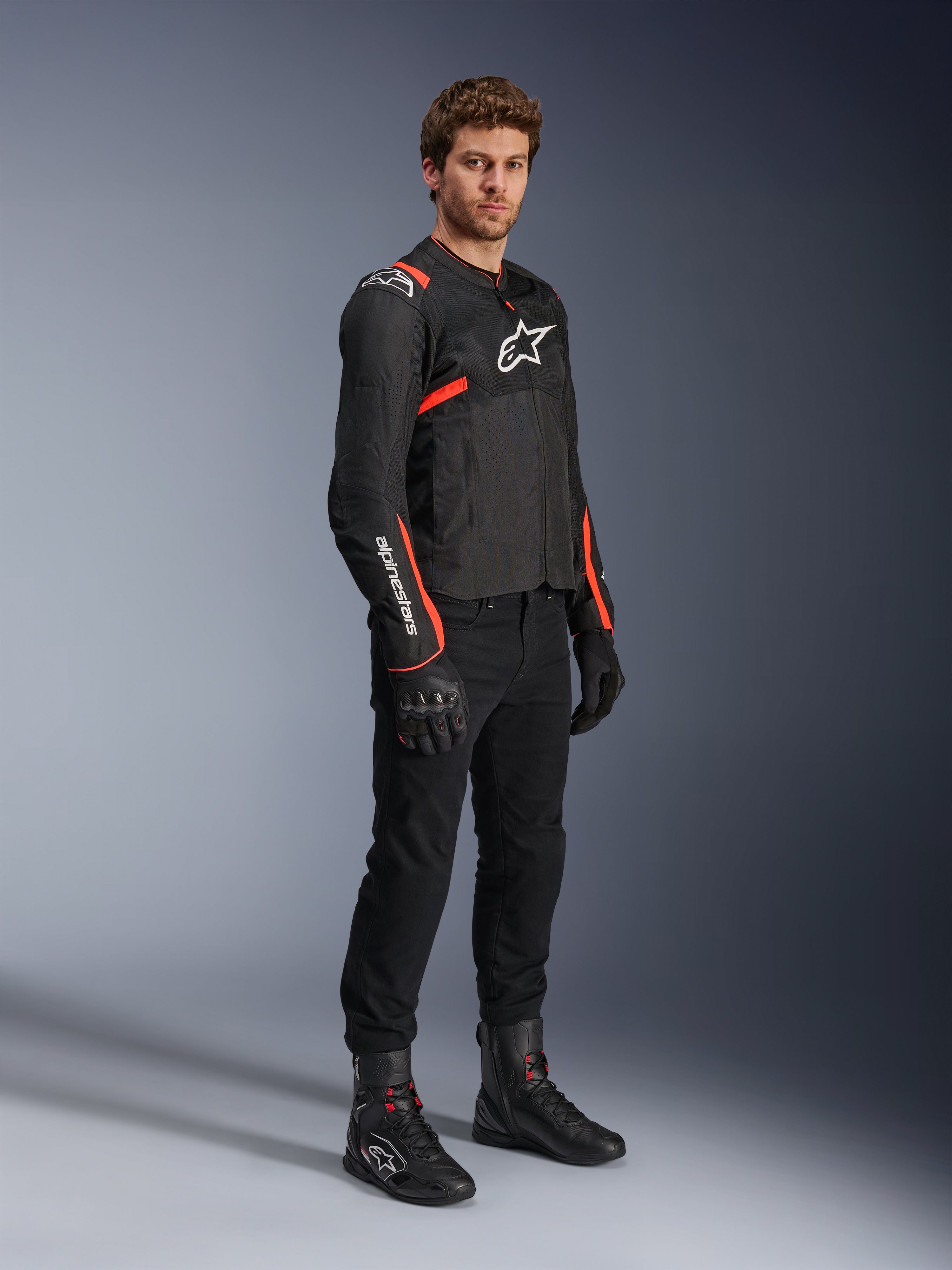 Modelo masculino a usar o Casaco Alpinestars T-Sps Air V2, preto com detalhes em vermelho fluorescente, combinado com calças de motociclismo pretas, luvas pretas e botas pretas, destacando o design desportivo com painéis de malha e logótipos Alpinestars brancos.