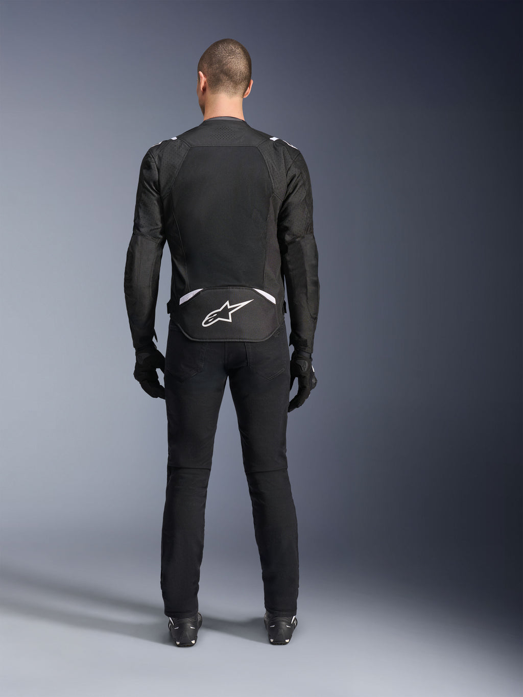 Casaco Alpinestars T-Sps Air V2, vista traseira, casaco de motociclismo em têxtil preto e branco, com um grande painel de malha nas costas para máximo fluxo de ar e logótipo Astars branco na parte inferior das costas, usado com calças e luvas de condução pretas
