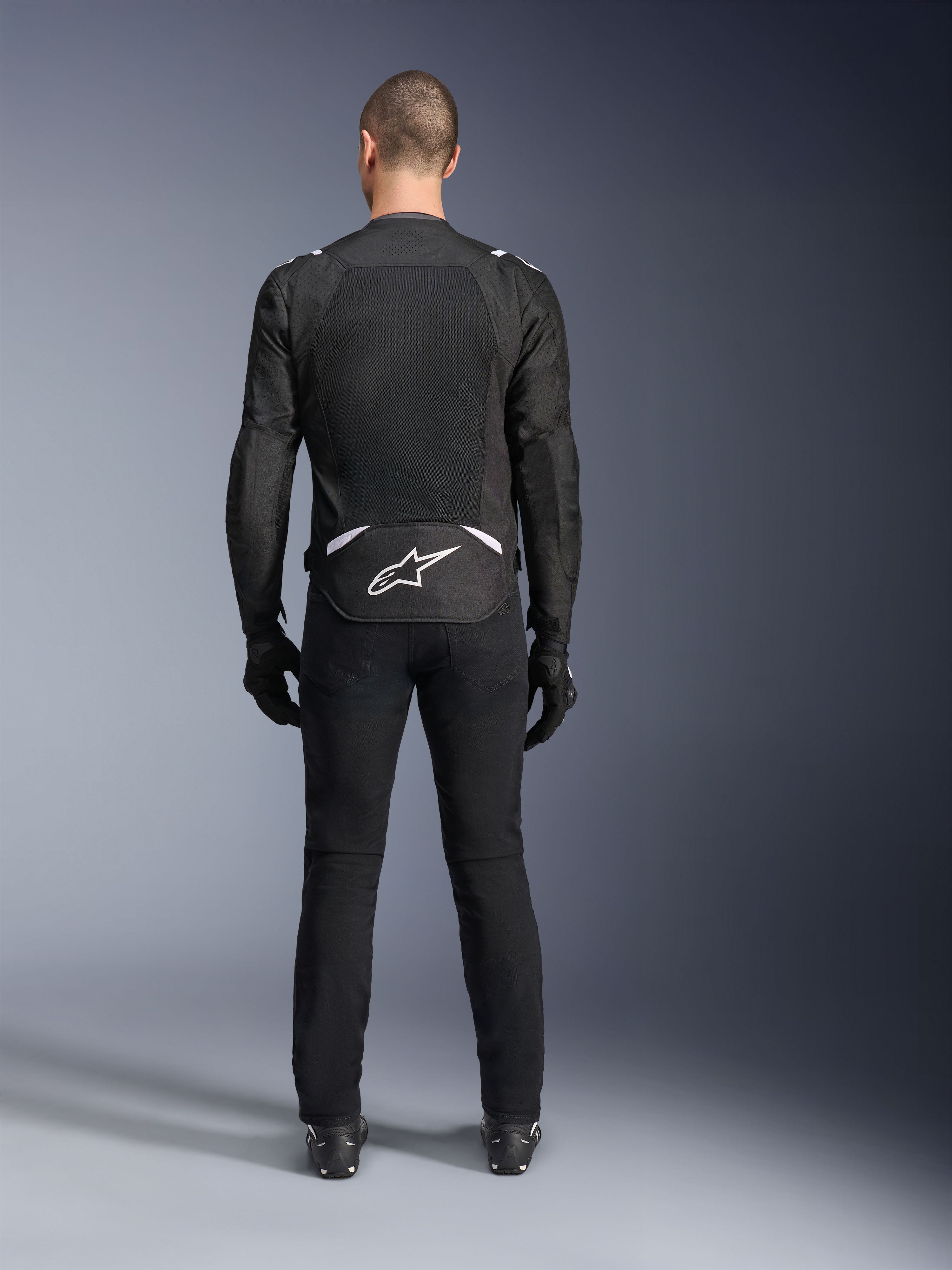 Casaco Alpinestars T-Sps Air V2, vista traseira, casaco de motociclismo em têxtil preto e branco, com um grande painel de malha nas costas para máximo fluxo de ar e logótipo Astars branco na parte inferior das costas, usado com calças e luvas de condução pretas
