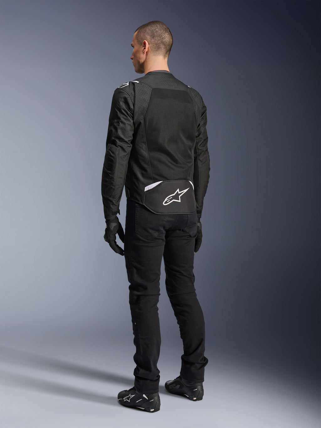 Casaco Alpinestars T-Sps Air V2, casaco de motociclismo em tecido, preto, vista traseira mostrando um grande painel de ventilação em malha e o logótipo Alpinestars branco na parte inferior das costas, combinado com calças e luvas de condução pretas para um visual urbano e desportivo.
