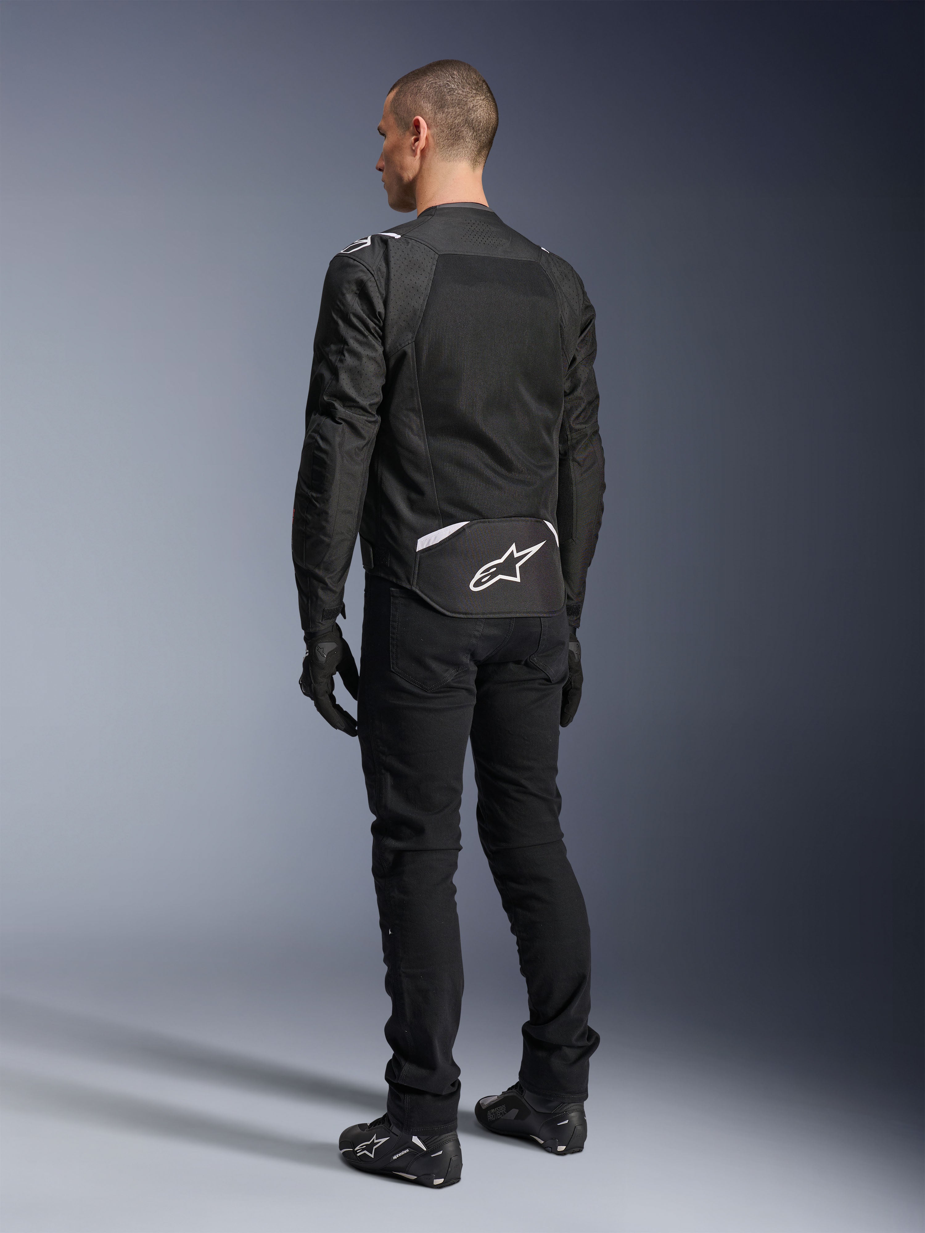 Casaco Alpinestars T-Sps Air V2, casaco de motociclismo em tecido, preto, vista traseira mostrando um grande painel de ventilação em malha e o logótipo Alpinestars branco na parte inferior das costas, combinado com calças e luvas de condução pretas para um visual urbano e desportivo.