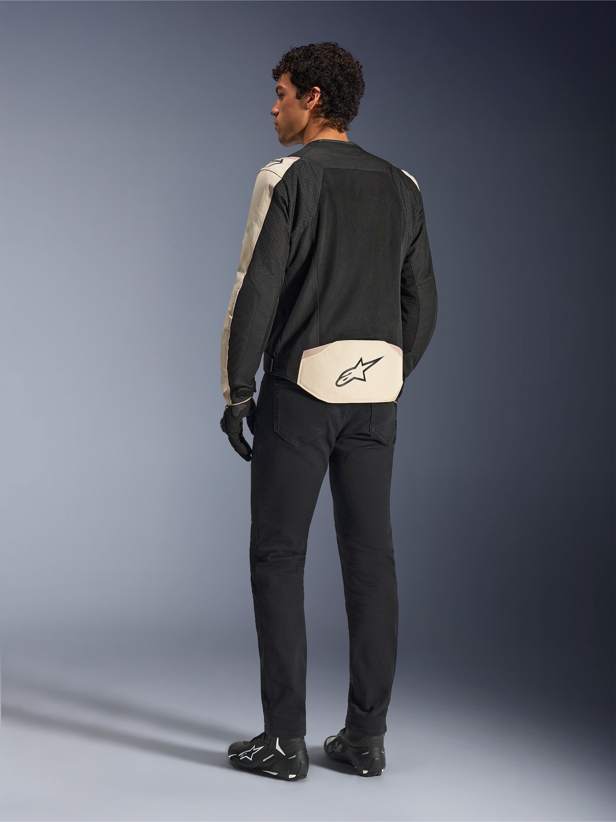 Casaco Alpinestars T-SPS Air V2, casaco têxtil para motociclista, Black Mastic Aluminum, vista traseira usado por modelo, com amplos painéis de malha técnica para fluxo de ar, detalhes em bege claro nas mangas e na zona lombar com o logótipo Alpinestars, combinado com calças e botas pretas.