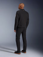 Casaco Alpinestars Bruiser, casaco de motociclismo urbano em preto, vista traseira num modelo, estilo clássico de vestuário de trabalho, com painéis de ombro pregueados e patch em espiral Oscar na manga, usado com calças pretas e sapatos pretos casuais com solas de borracha natural.