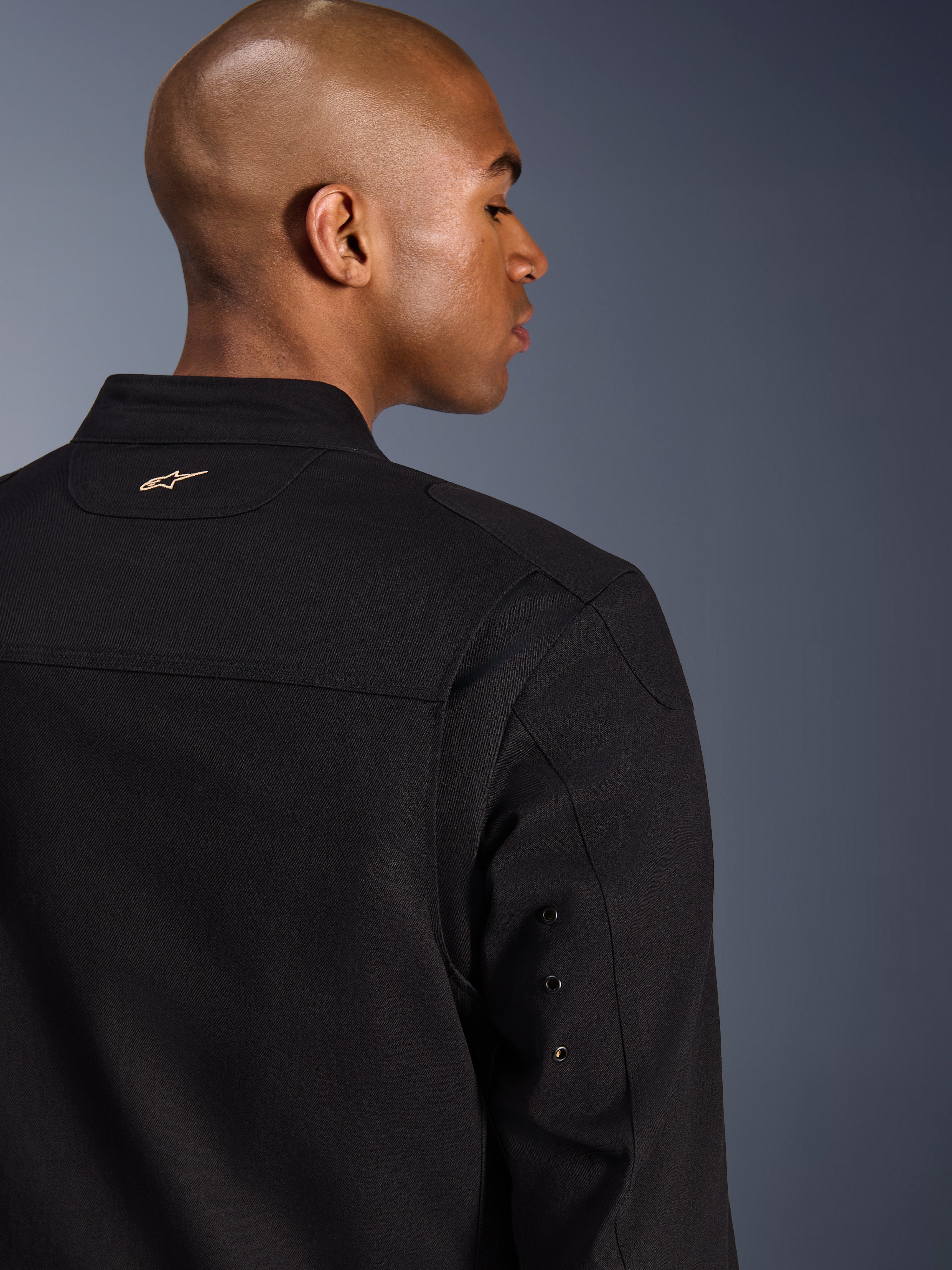 Alpinestars Bruiser Jacket, casaco de motociclismo urbano em tecido, preto, pormenor da vista traseira com logótipo dourado bordado na parte superior das costas, detalhes de ilhós metálicos nas mangas e painéis de ombros pregueados para maior mobilidade