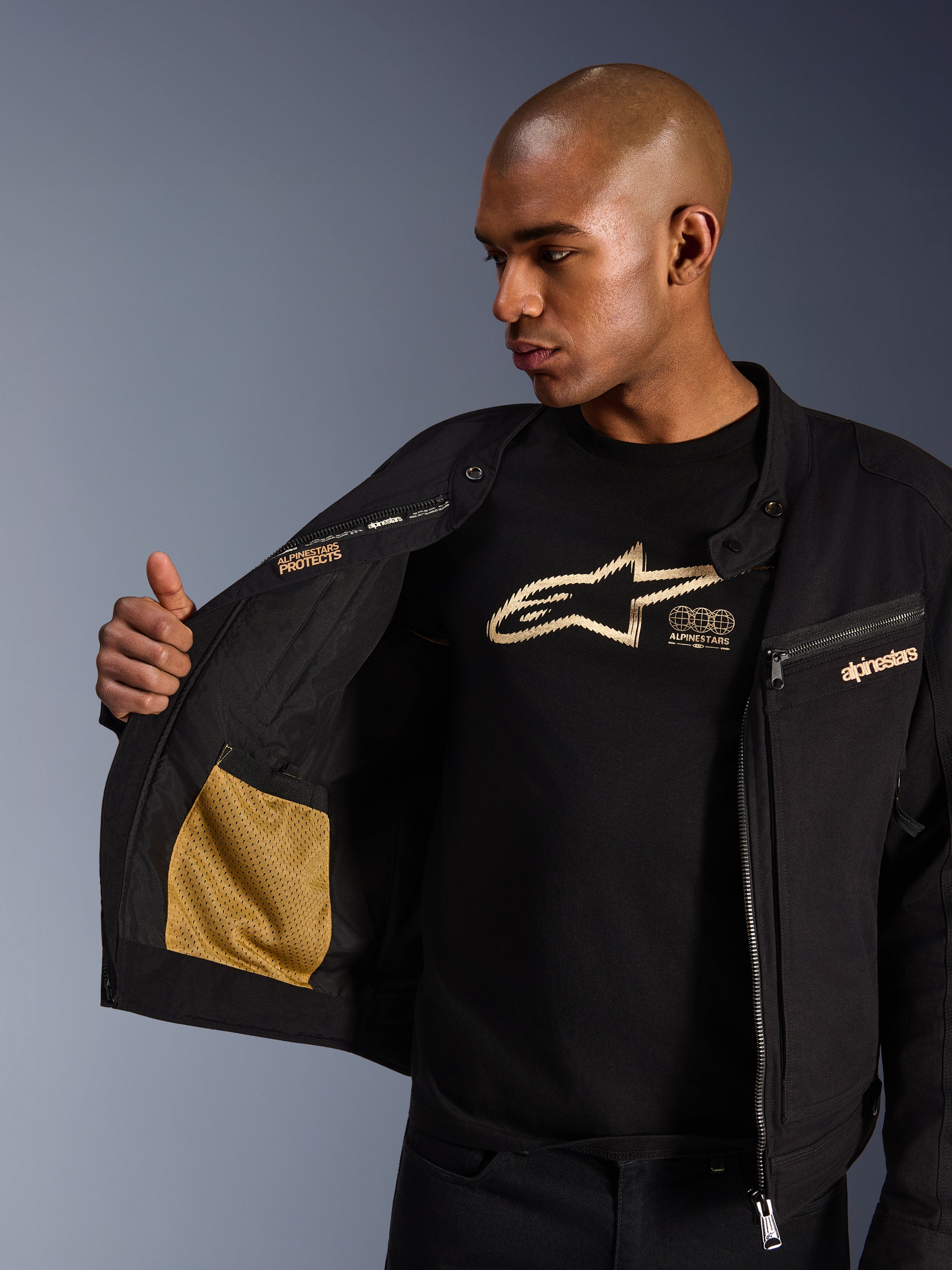 Alpinestars Bruiser Jacket, casaco de motociclismo urbano preto, vista aproximada mostrando o interior aberto com bolso em rede dourada e marca 'Alpinestars Protects', usado sobre uma t-shirt preta com logótipo, equipamento têxtil com estética de vestuário de trabalho