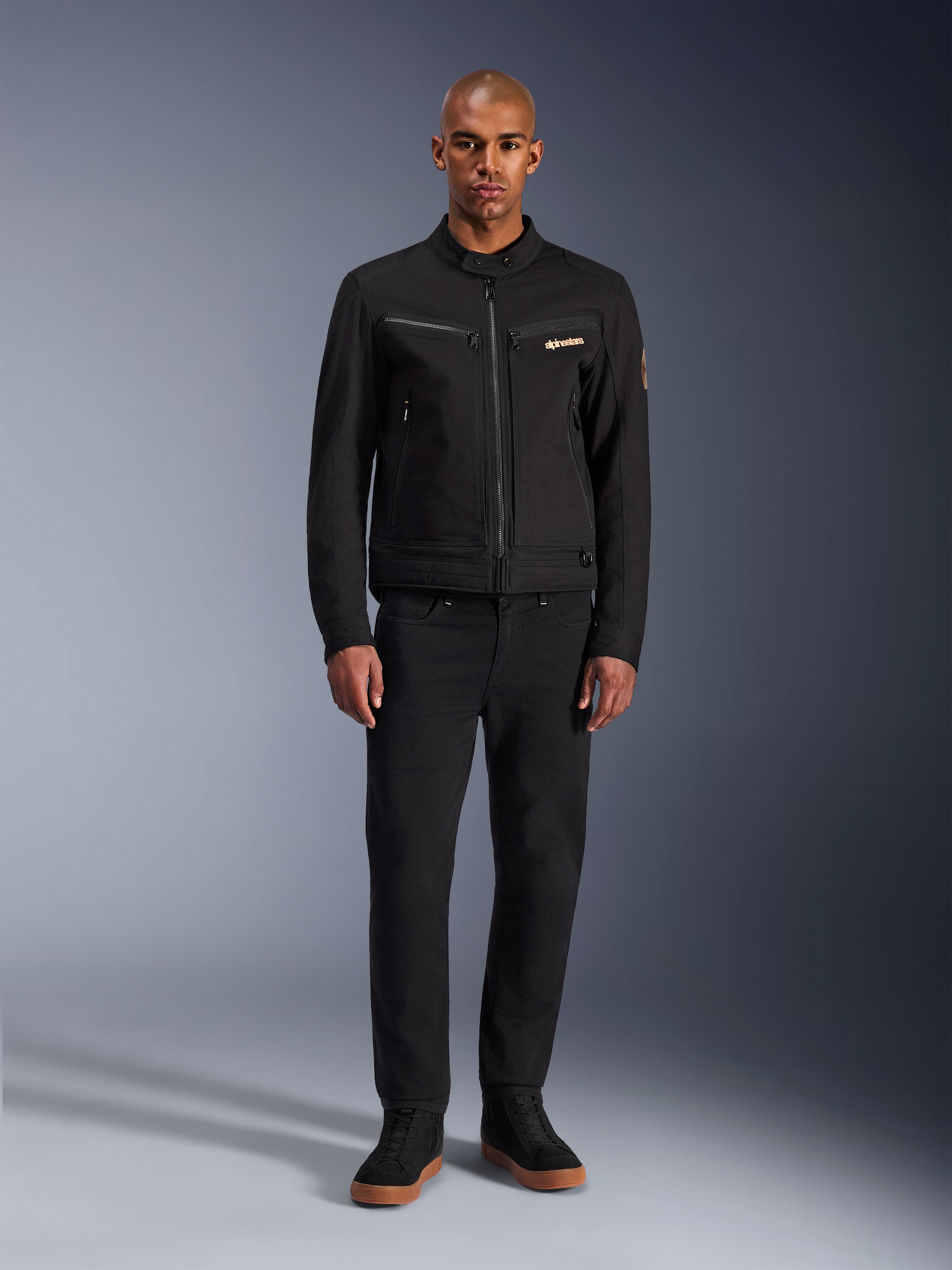 Casaco Alpinestars Bruiser, casaco de motociclismo urbano preto, usado por modelo com calças pretas e sapatilhas de cano alto, vista frontal com bolsos de peito com fecho e logótipo bordado, design clássico inspirado em vestuário de trabalho para deslocações quotidianas
