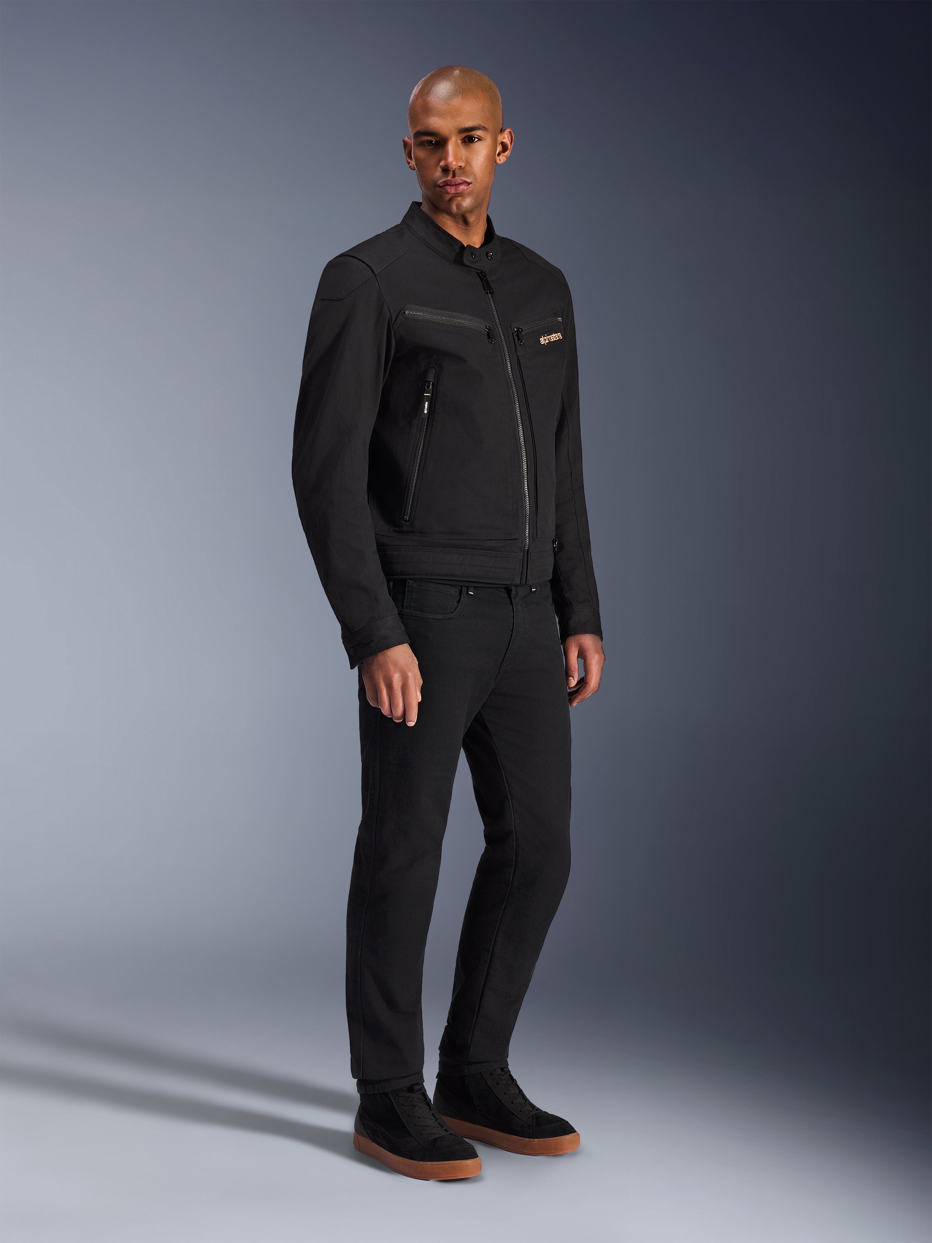 Alpinestars Bruiser Jacket, casaco de motociclismo urbano, preto, vestido por um modelo com calças pretas e sapatilhas, com bolsos no peito com fecho de correr, tecido em sarja de policotton e um design elegante inspirado no vestuário de trabalho para condução urbana