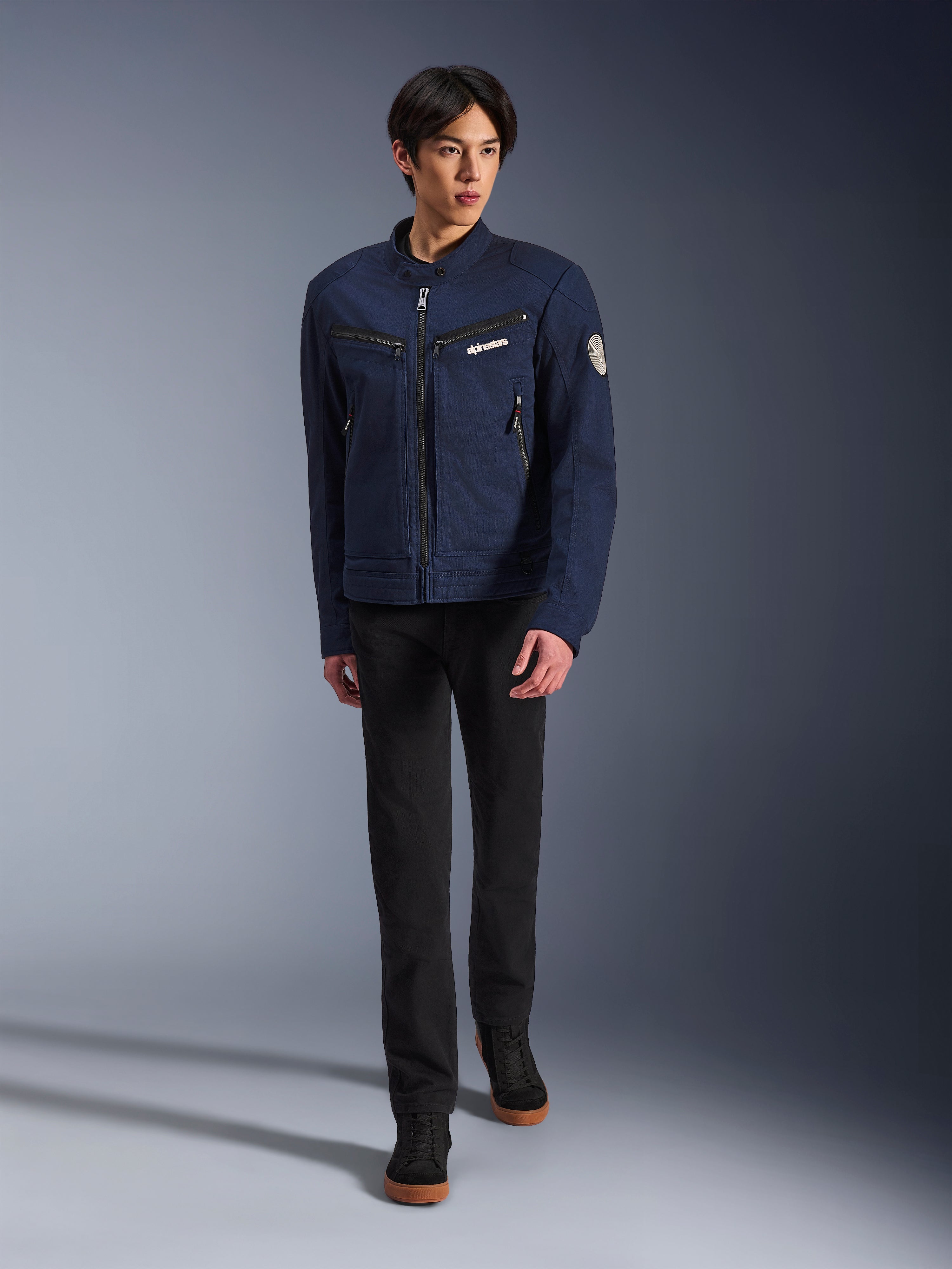 Male model wearing Alpinestars Bruiser Jacket in Dark Blue, urban textile motorcycle jacket, paired with black pants and black sneakers, workwear-inspired aesthetic with zippered pockets and circular sleeve patch, contemporary streetwear for motorcycle riding.