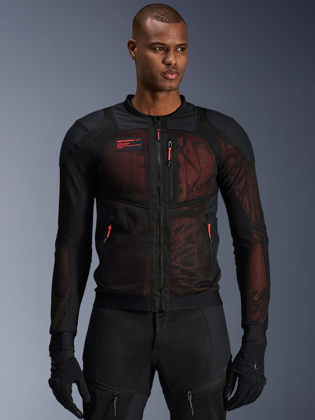 Casaco Alpinestars Troop-Air Pro, casaco de motociclismo técnico em malha blindada em preto e vermelho fluo, vestido por um modelo, com grandes painéis de ventilação em malha, camada interior vermelha e puxadores de fecho vermelhos, estilo de condução urbano.