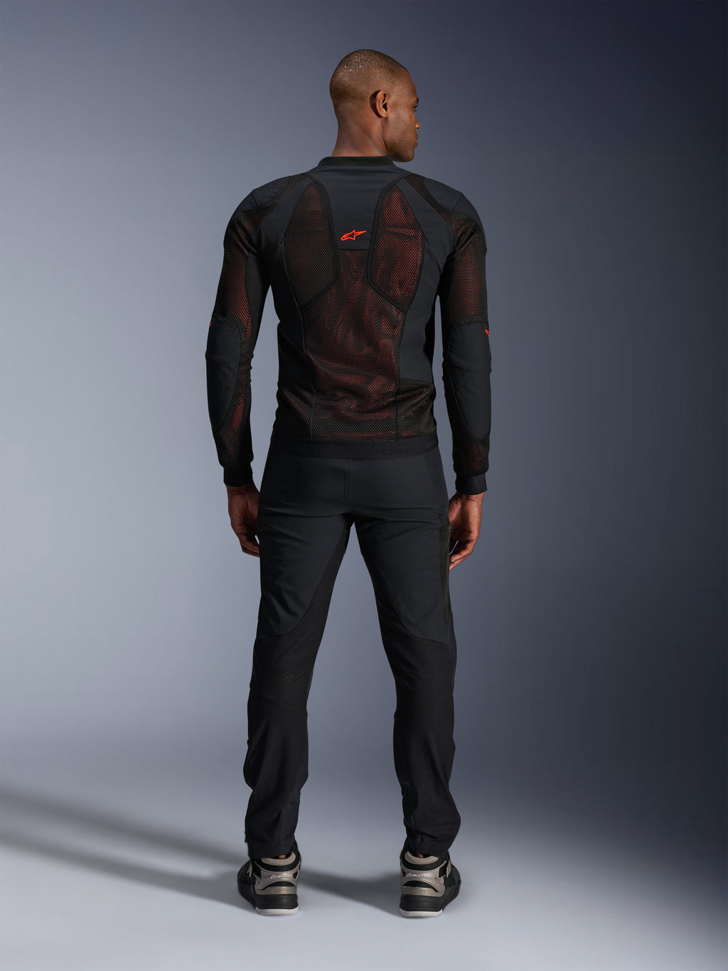 Casaco Alpinestars Troop-Air Pro em Preto e Vermelho Fluo, vista traseira no modelo, casaco de rede técnico com proteções e painéis respiráveis, logótipo vermelho na parte superior das costas, usado com calças pretas e sapatilhas, vestuário de motociclismo urbano