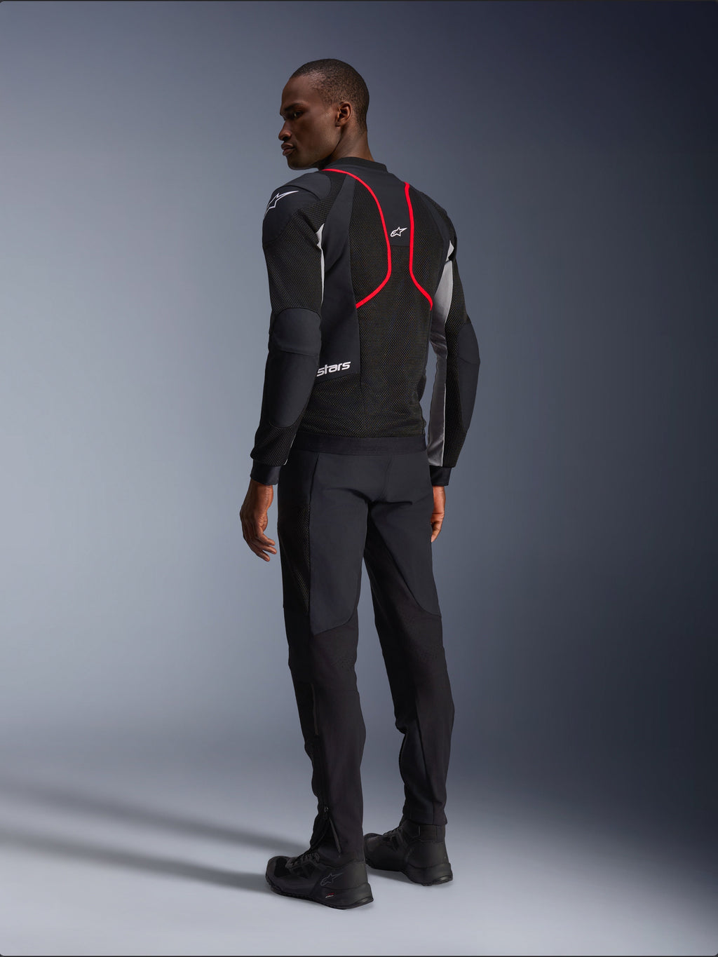 Alpinestars Troop-Air Pro Jacket, casaco de motociclismo, preto, cinzento e vermelho vivo, vista de costas num modelo, com linhas de realce vermelhas e painéis de ventilação em malha, usado com calças e botas pretas, estilo de motociclismo urbano