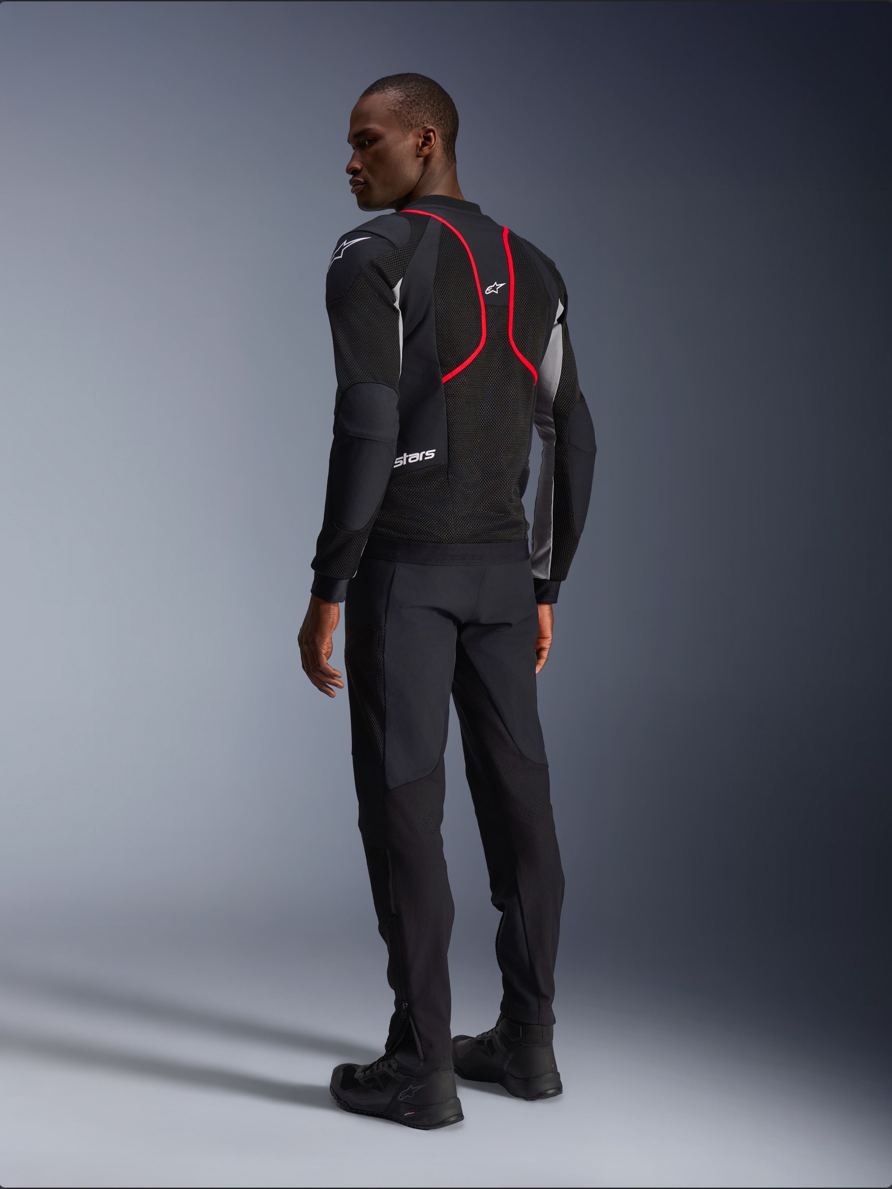 Alpinestars Troop-Air Pro Jacket, casaco de motociclismo, preto, cinzento e vermelho vivo, vista de costas num modelo, com linhas de realce vermelhas e painéis de ventilação em malha, usado com calças e botas pretas, estilo de motociclismo urbano