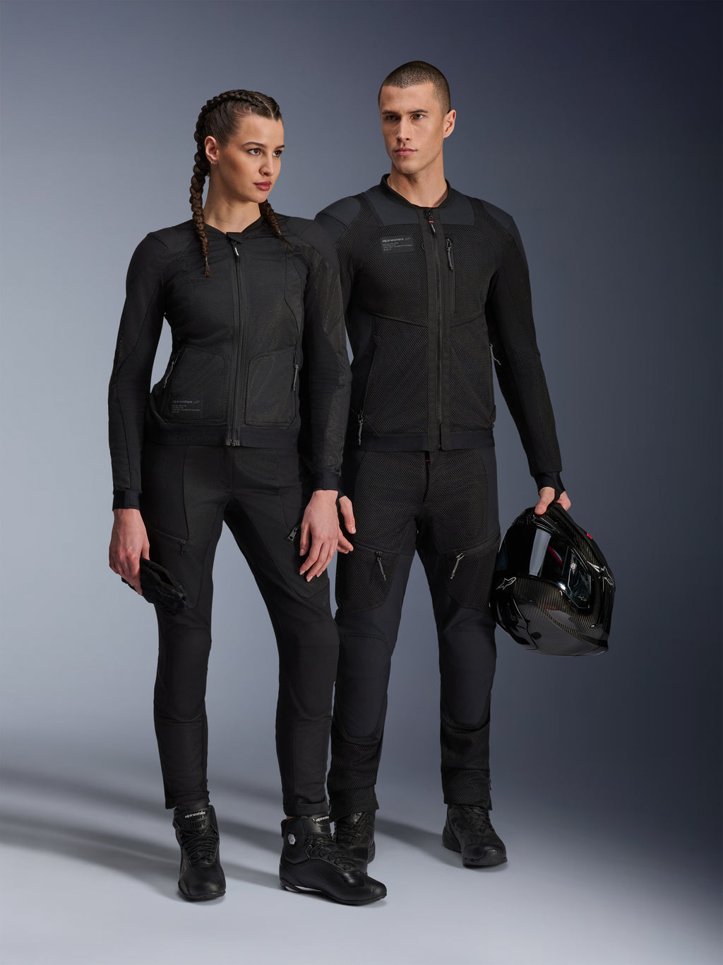 Alpinestars Stella Troop-Air Jacket, Man and woman wearing matching all-black technical motorcycle gear, featuring mesh construction for airflow, black pants and boots, man holding a black helmet, urban riding apparel.