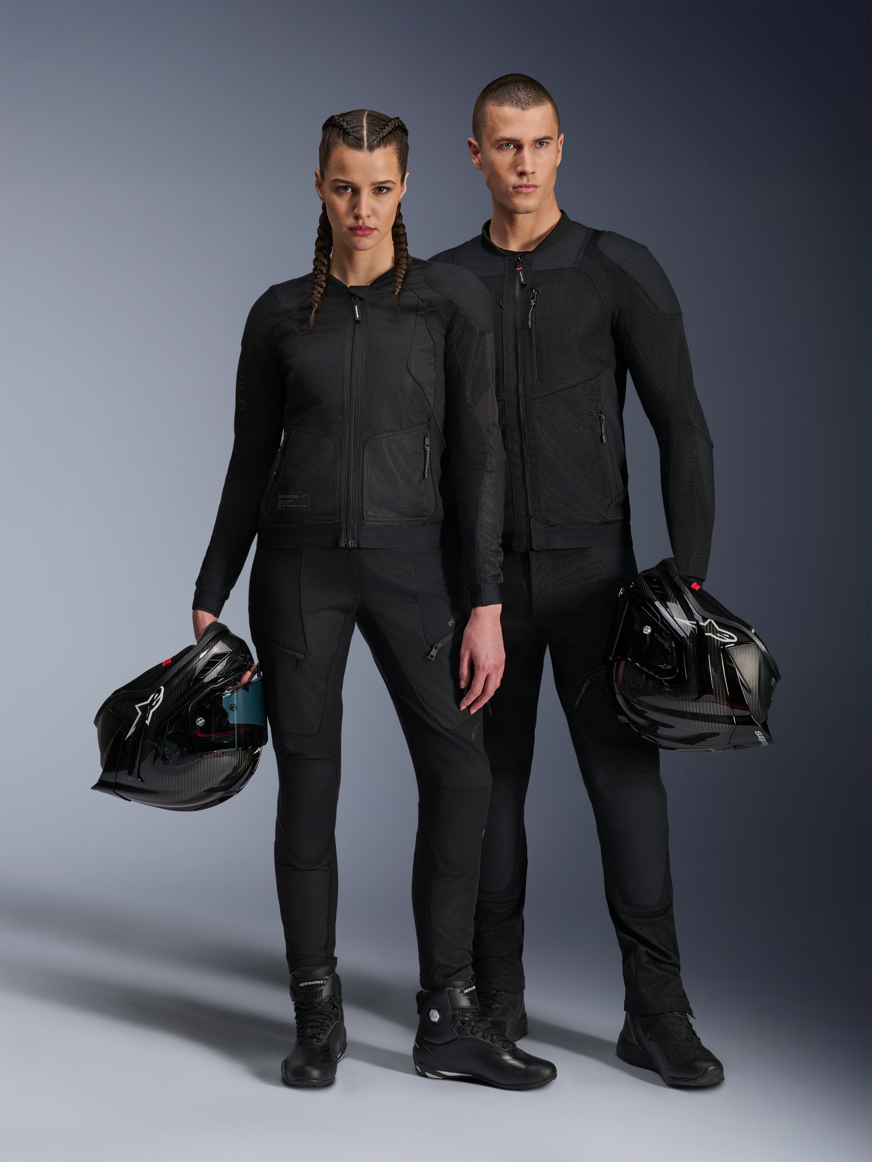Modelos masculino e feminino vestindo casacos Alpinestars Troop-Air Pro em preto, equipamento técnico de condução em malha blindada, combinados com calças e botas pretas a condizer, cada um segurando um capacete preto, exibindo vestuário urbano coordenado para motociclismo.